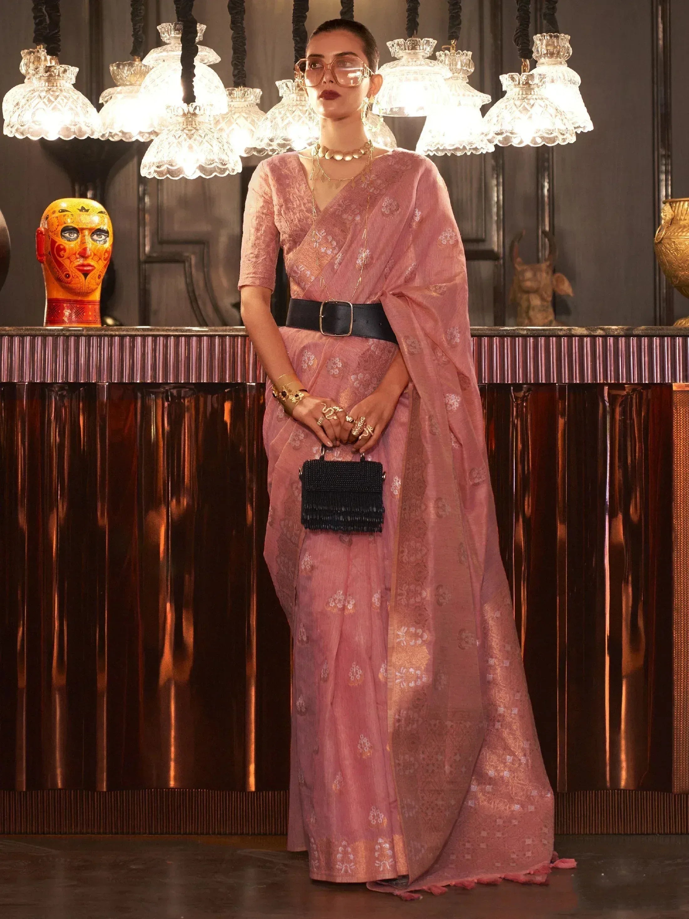 Pink floral saree with black belt, styled with gold jewelry and clutch, under crystal chandeliers
