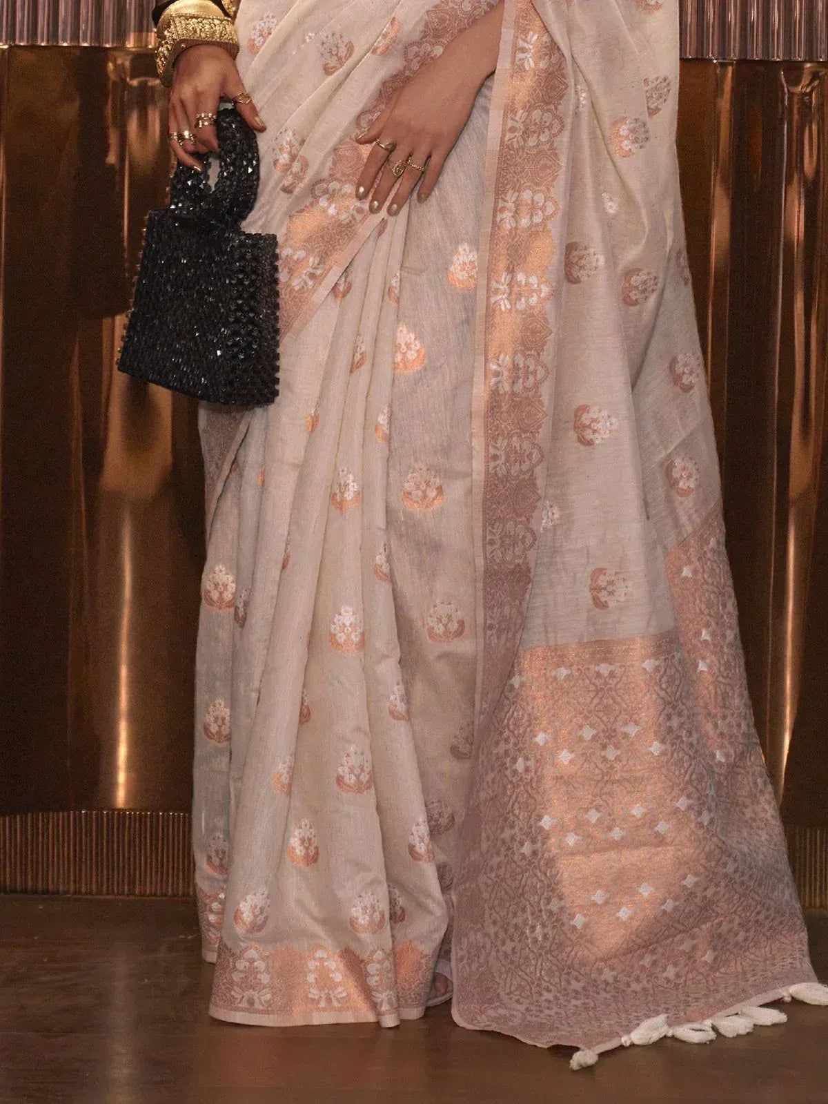 Elegant beige saree with peach floral motifs and intricate border, paired with black embellished clutch for ethnic wear.