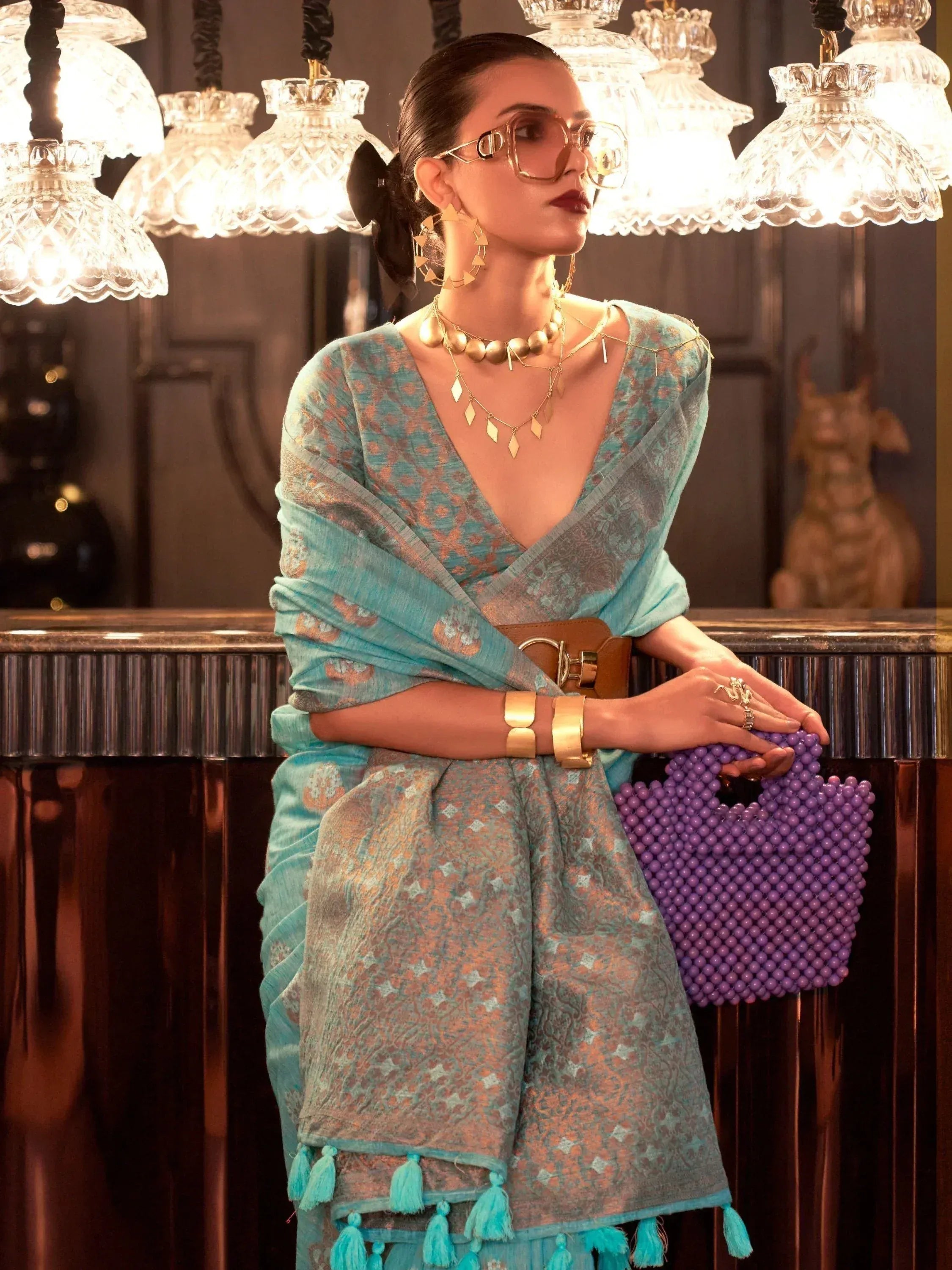 Stylish woman in teal printed ethnic gown with tassels, gold jewelry, and purple beaded clutch under chandelier
