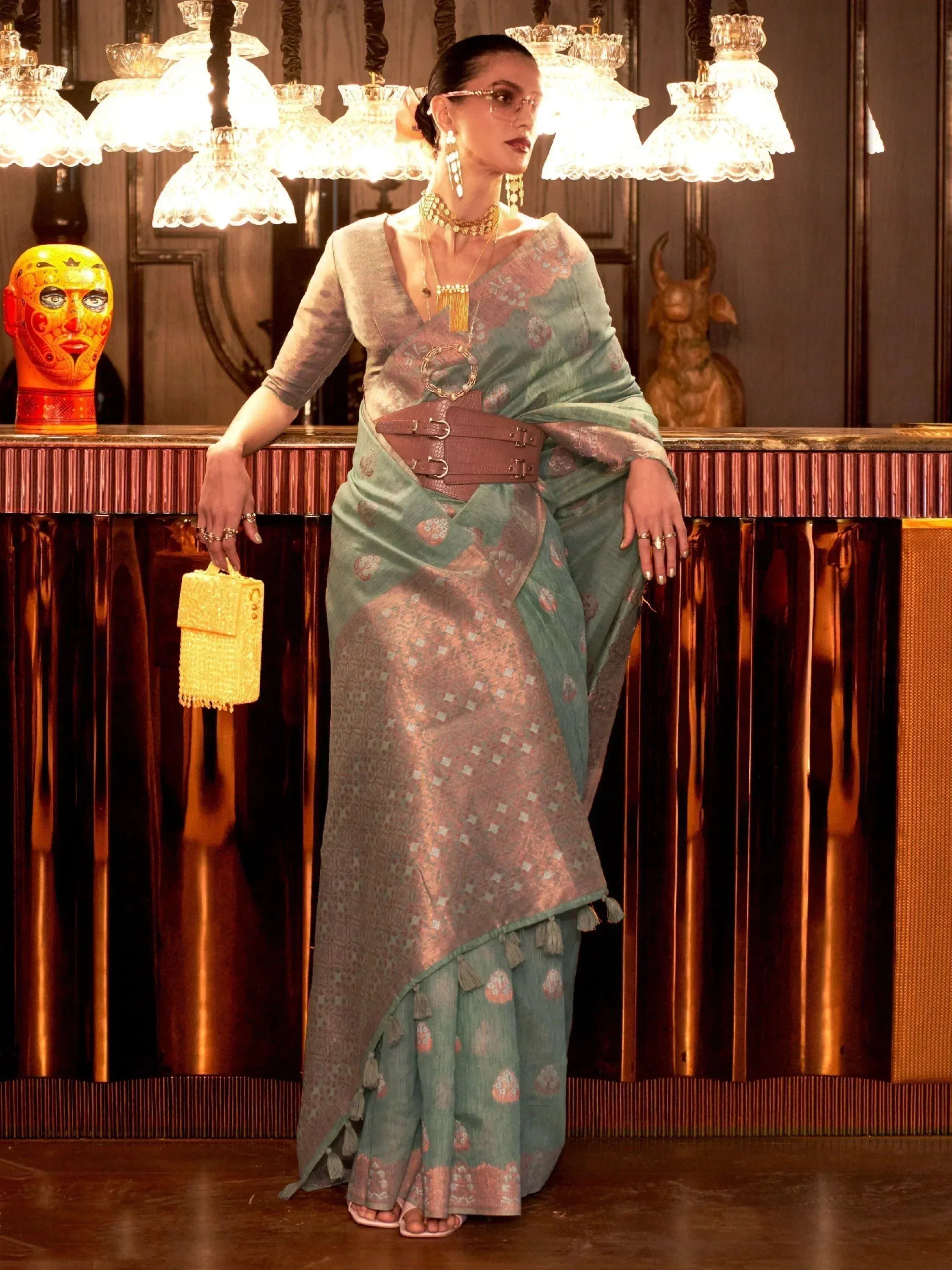 Stylish woman in designer saree with gold embroidery, tassels, and wide belt, holding yellow clutch under chandelier.