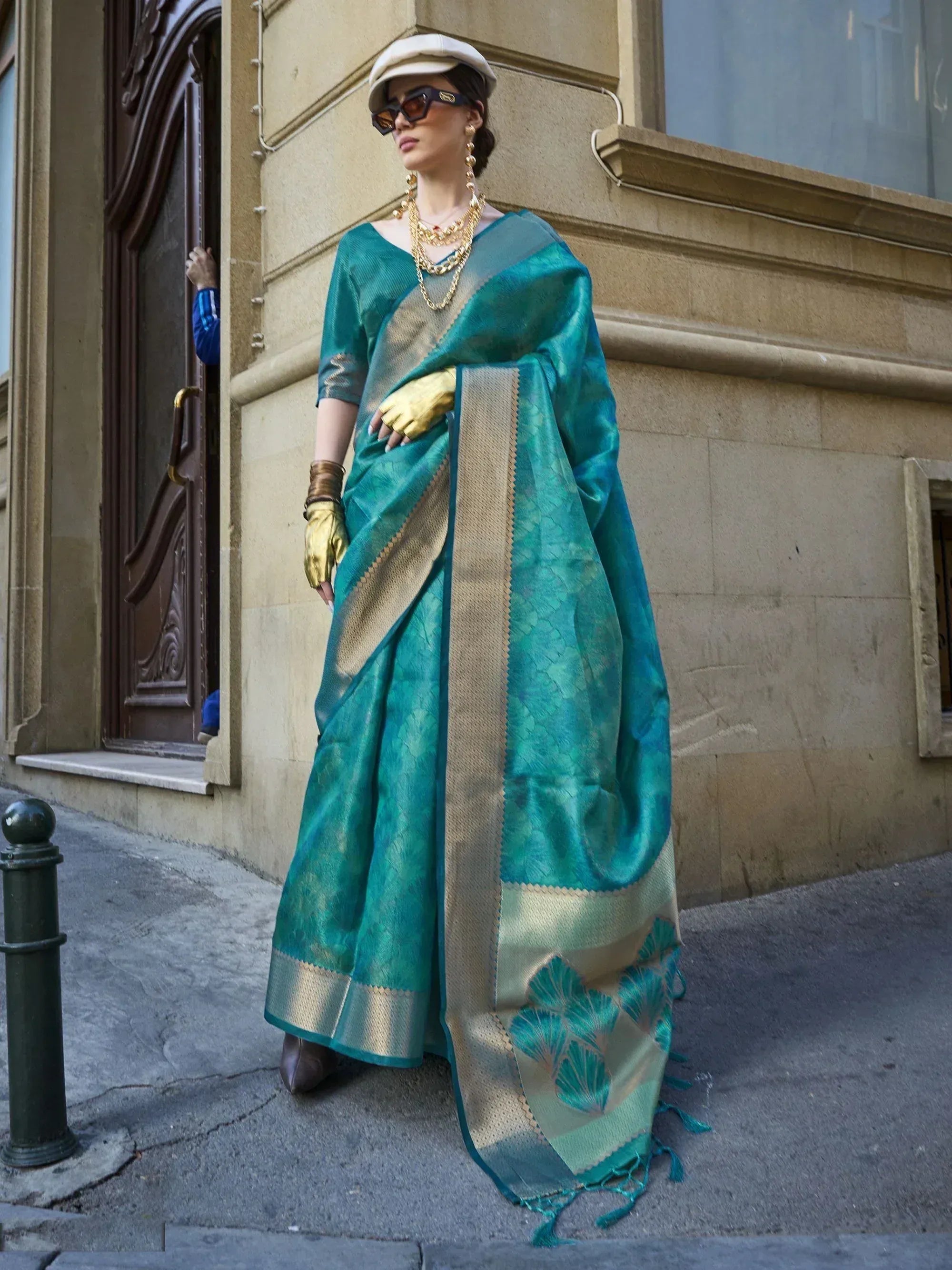Teal silk saree with gold border and leaf motifs, styled with cap and sunglasses for ethnic fashion