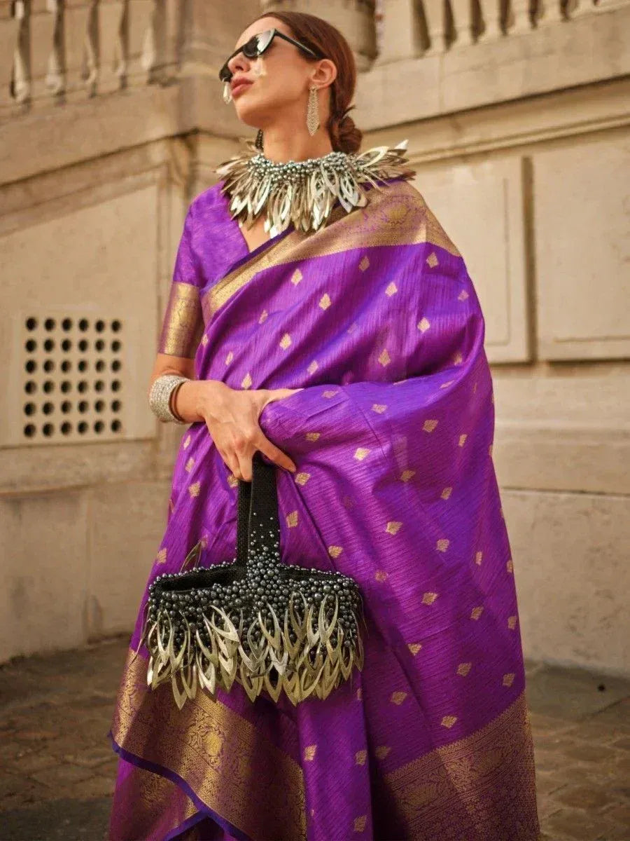 Purple designer saree with gold motifs, statement necklace, and embellished handbag for ethnic wear