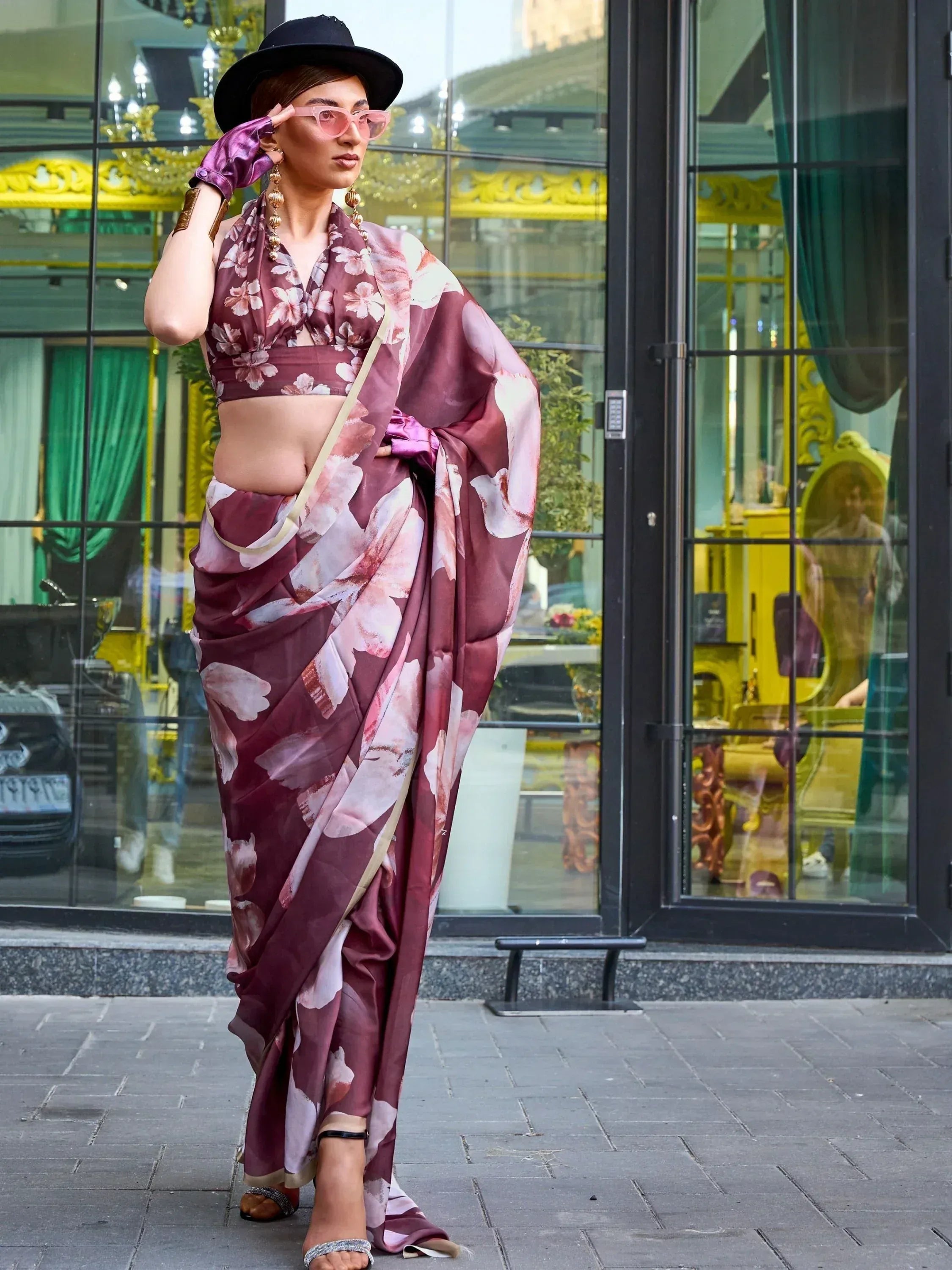 Woman in floral print saree with crop top, black hat, and pink sunglasses, styled for modern ethnic fashion.