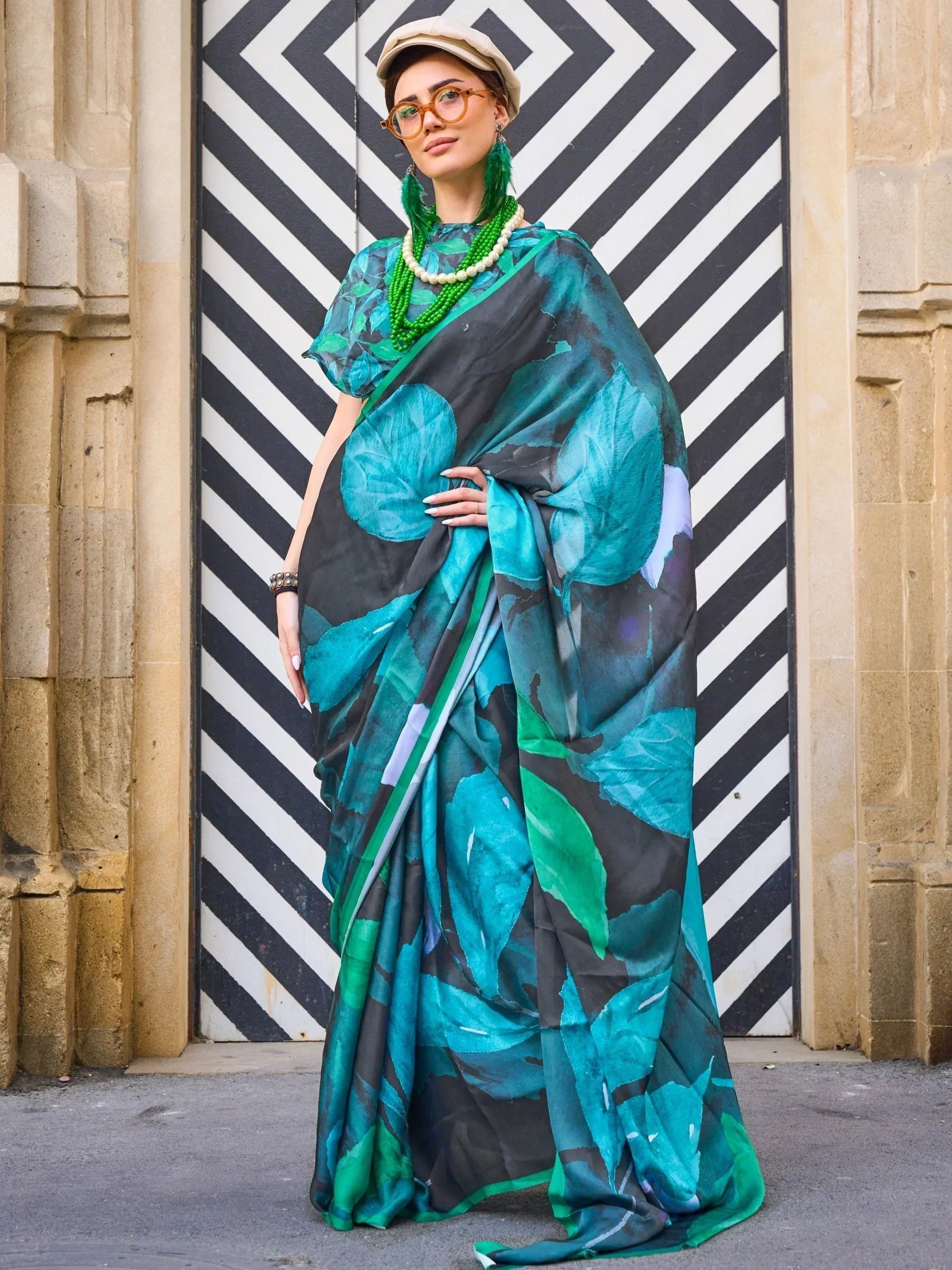Woman in stylish teal and green leaf print saree with matching blouse, modern cap, and statement jewelry.