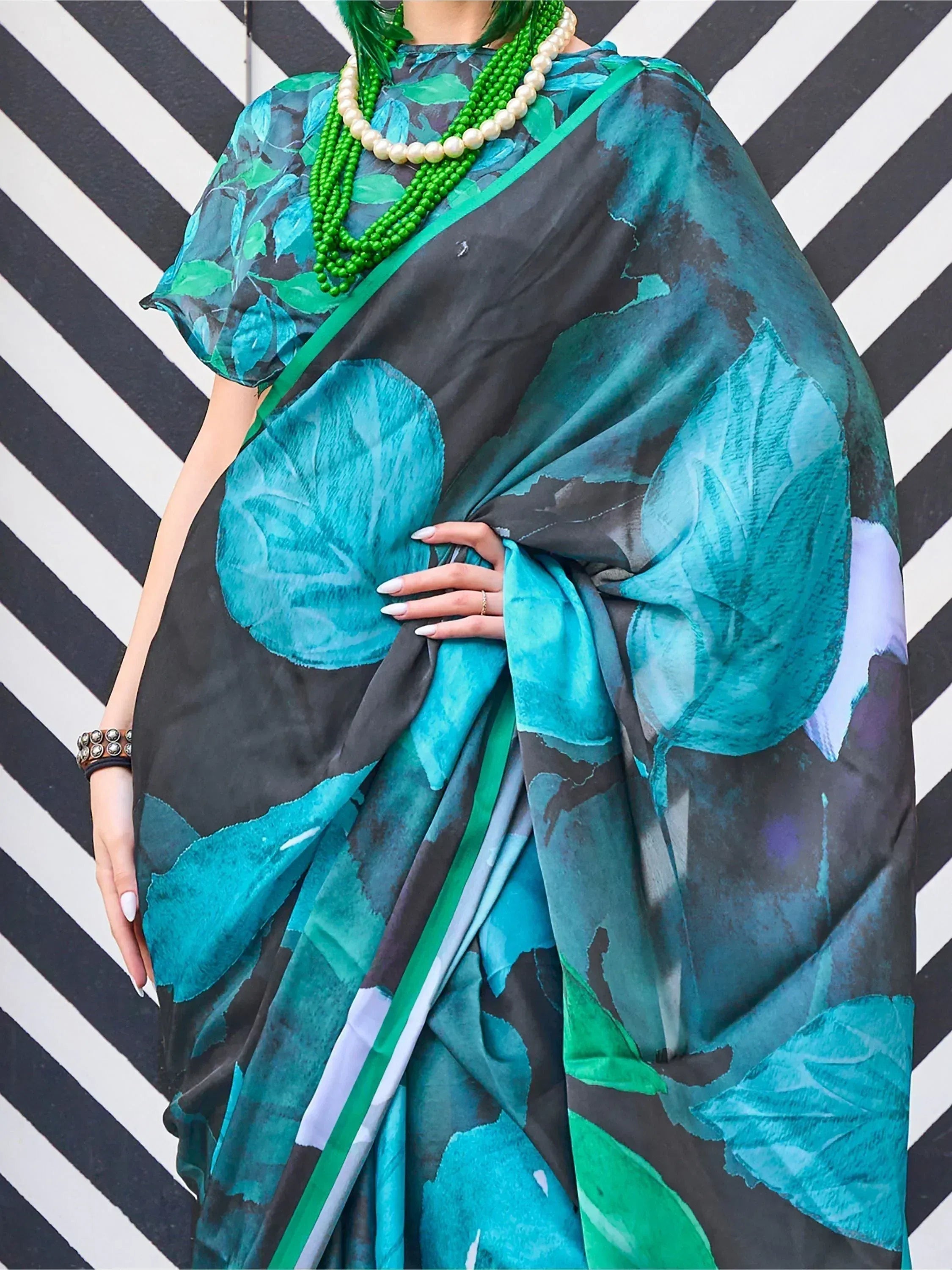 Teal and black floral print saree with green border, styled with pearl and green bead necklaces against striped backdrop.