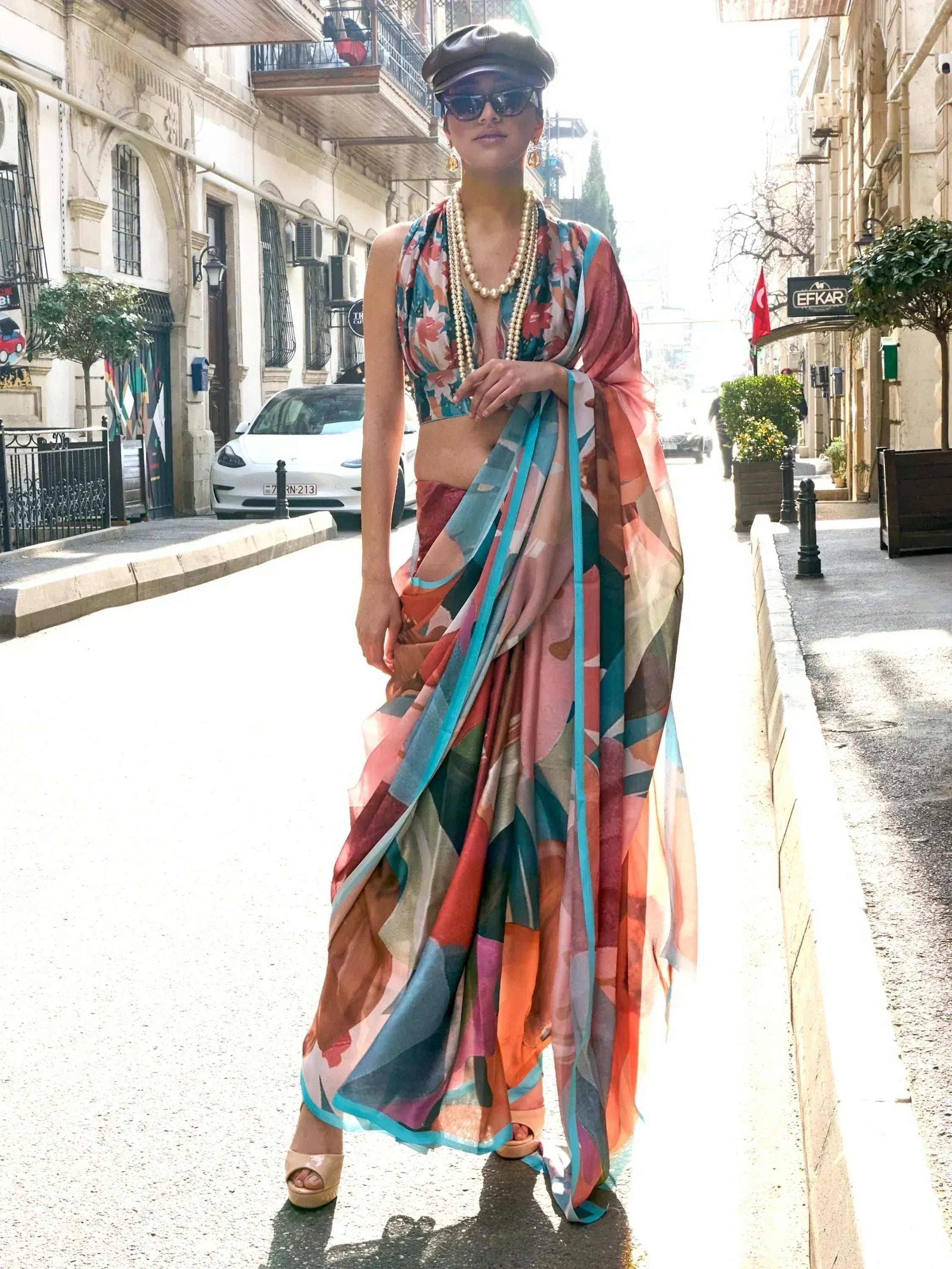 Woman in colorful floral saree with crop top, pearl necklace, and cap walking on city street