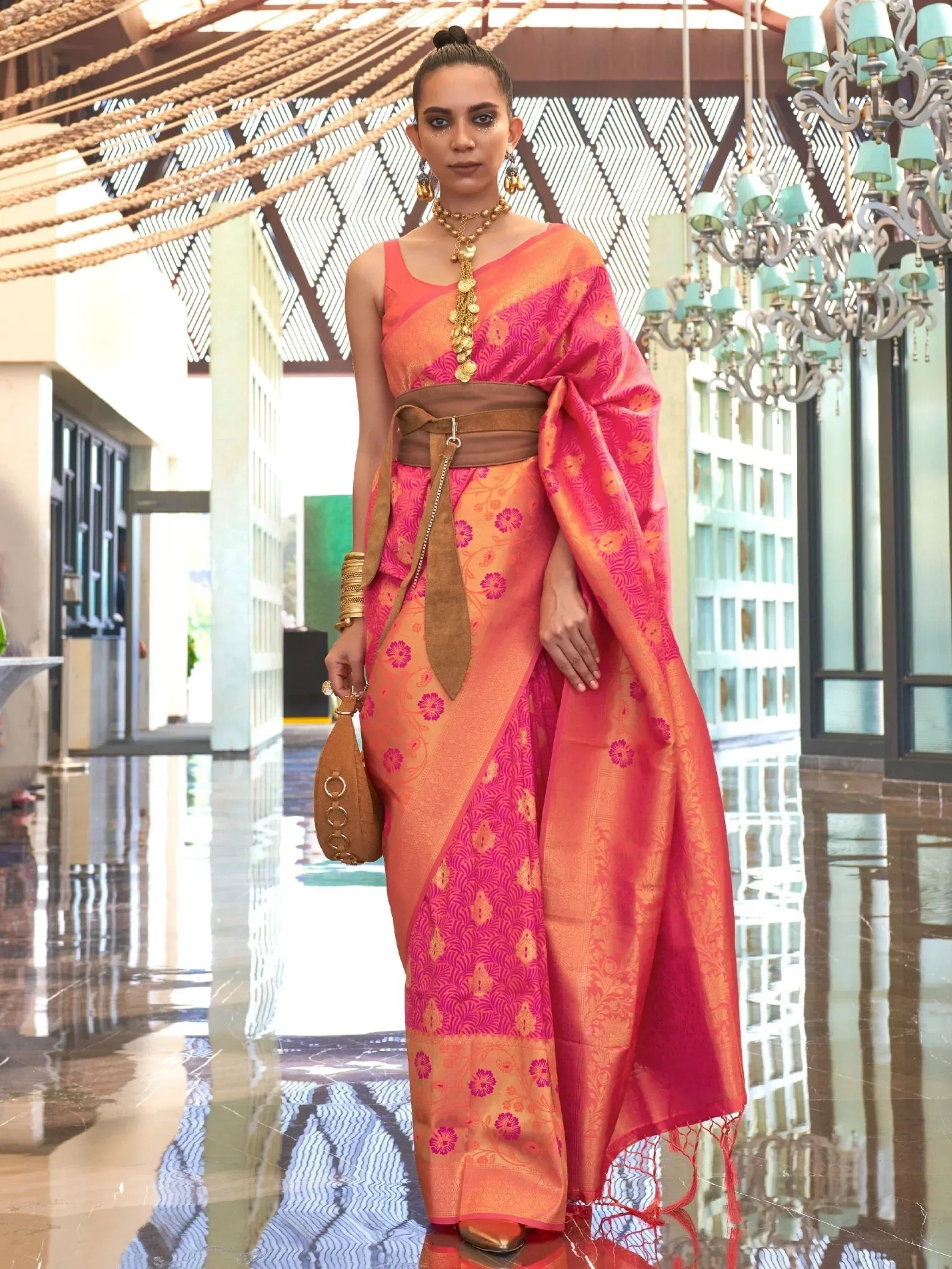 Woman in vibrant pink and orange designer saree with floral motifs, gold jewelry, and brown belt, styled for ethnic occasions.