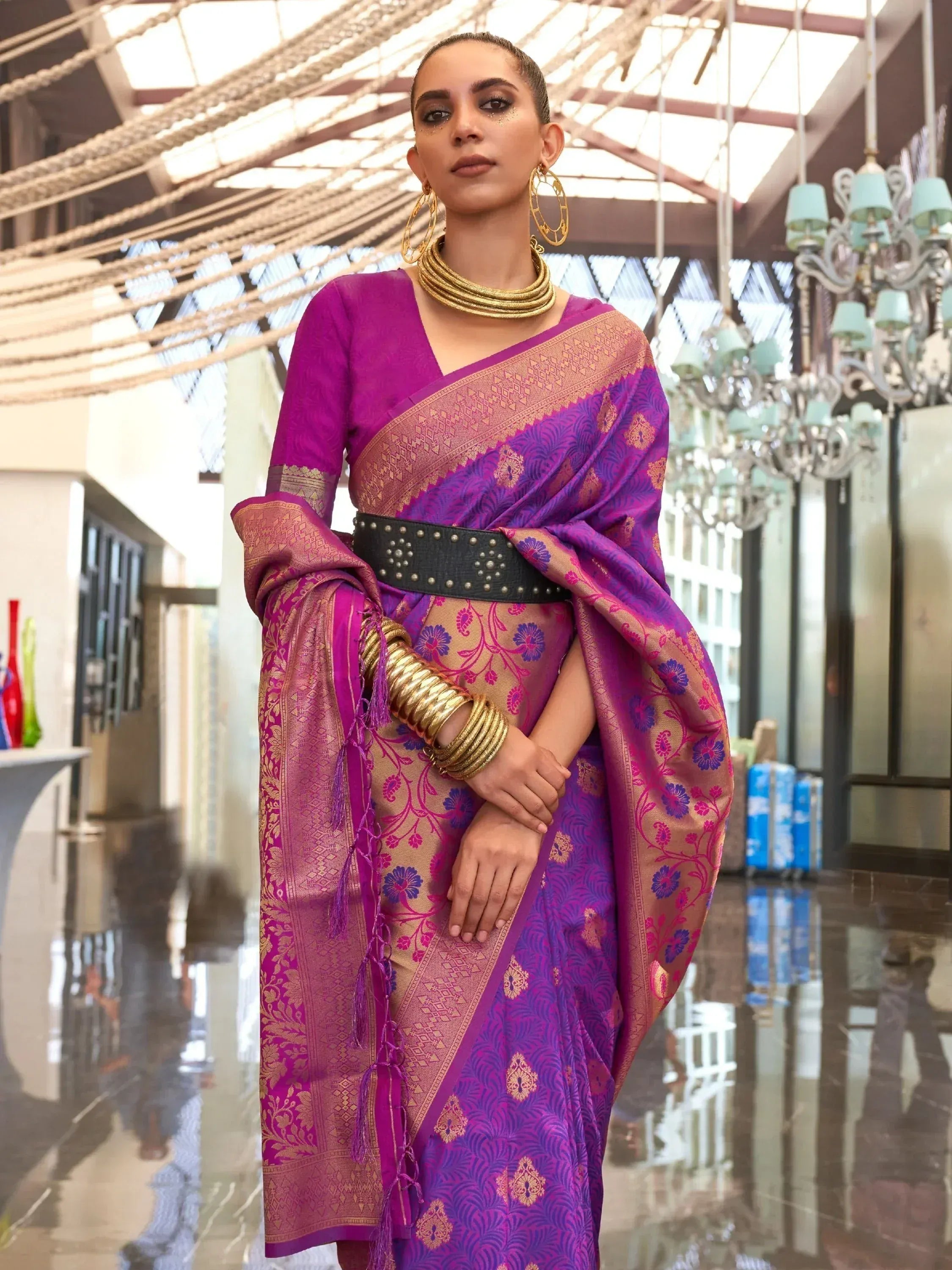 Purple and gold designer saree with floral motifs, paired with statement gold jewelry and a black studded belt.