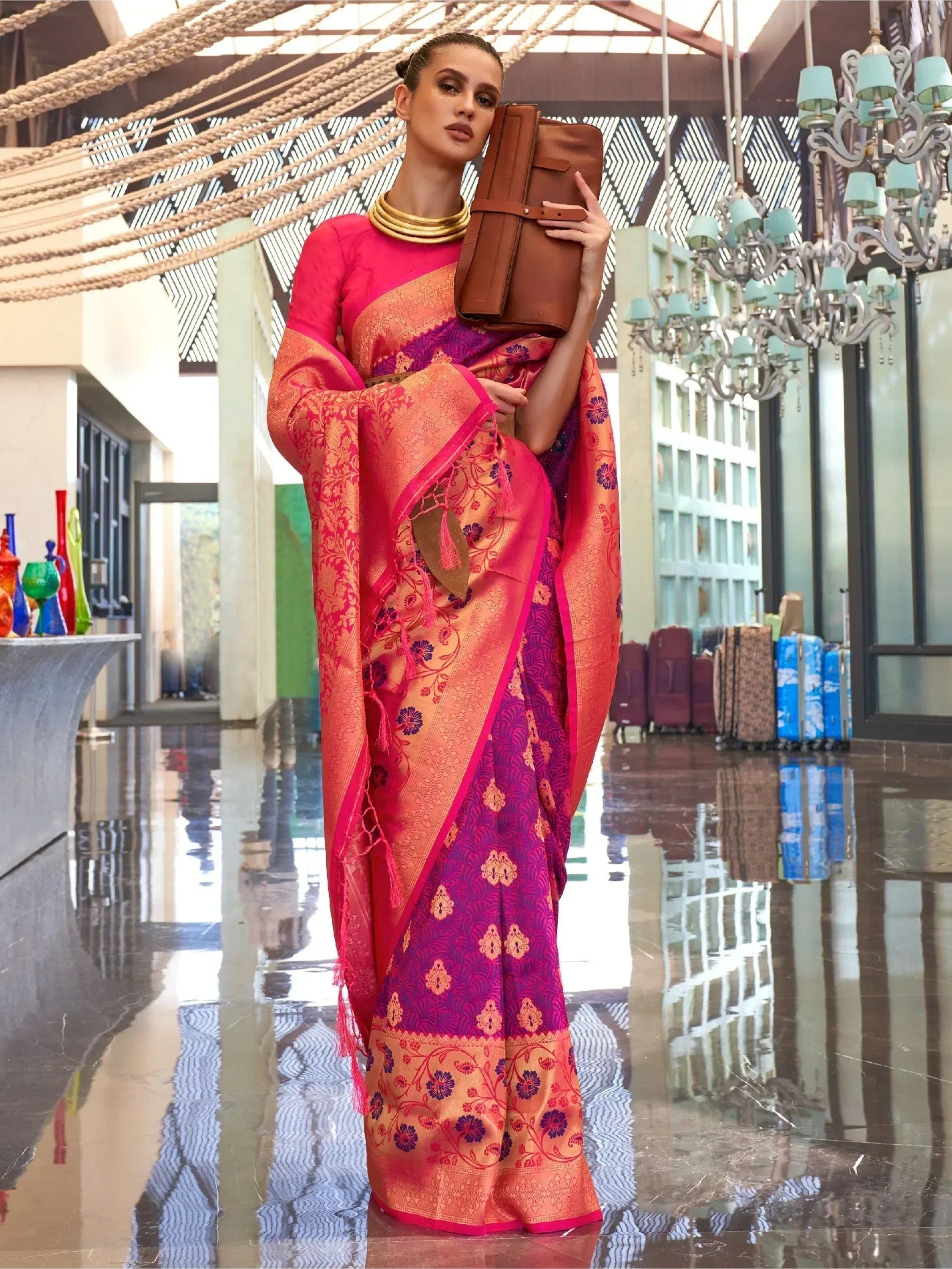 Woman in vibrant pink and purple designer saree with gold embroidery, holding brown clutch in luxury lobby