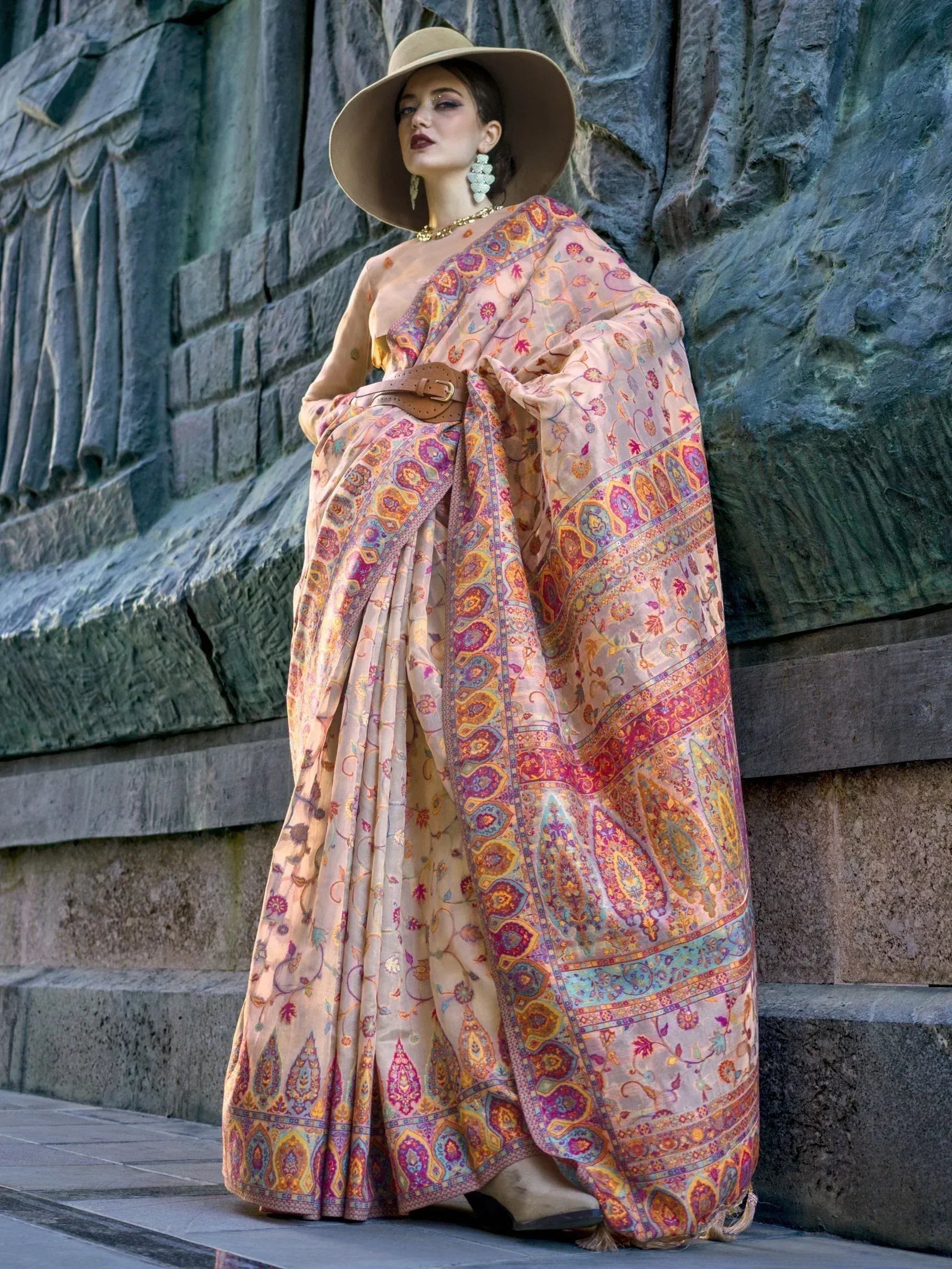Woman in floral printed saree with wide hat, standing by stone wall, ethnic Indian wear