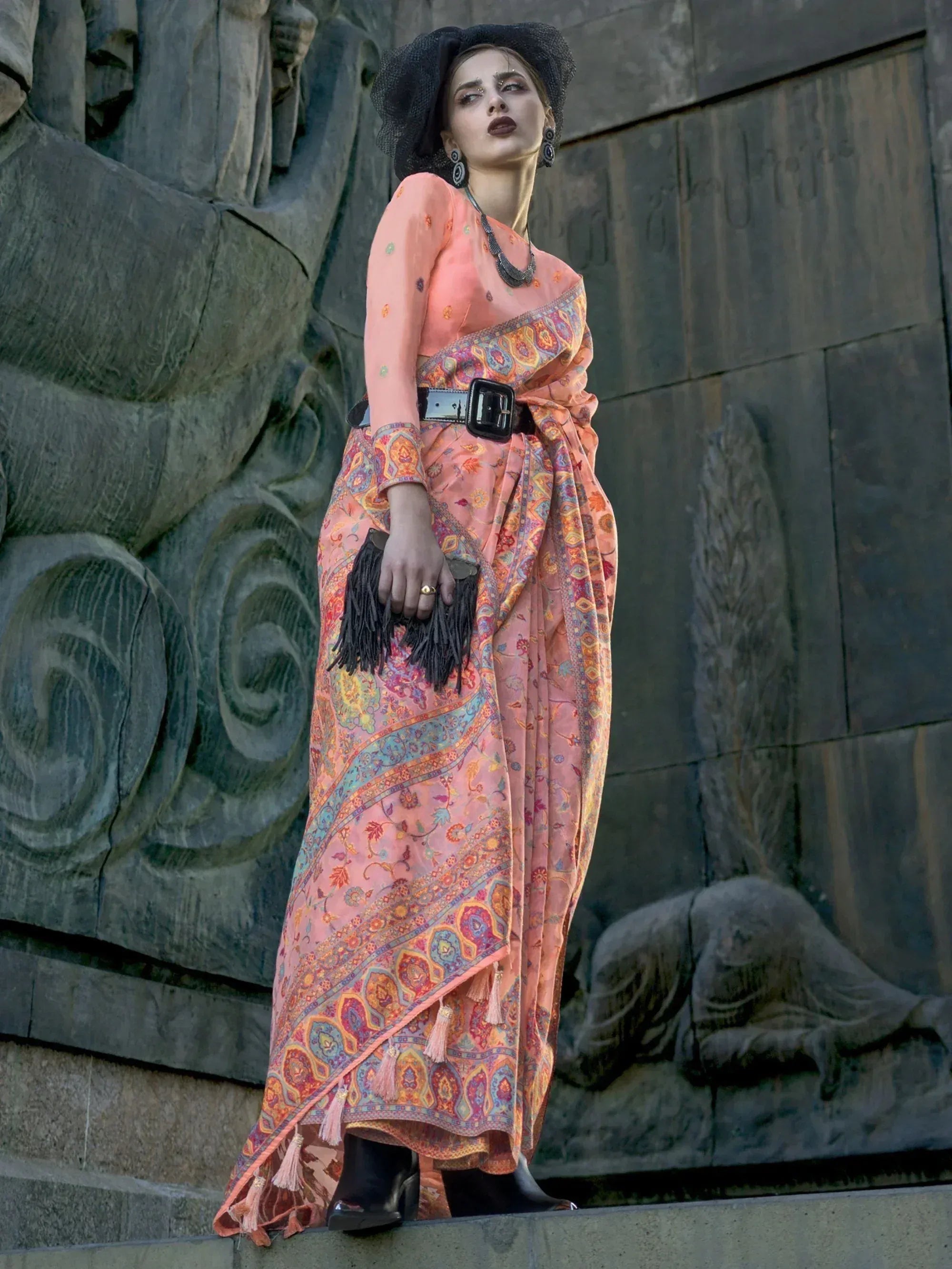Woman in peach printed saree with belt, black hat, and fringe clutch, styled for ethnic fashion
