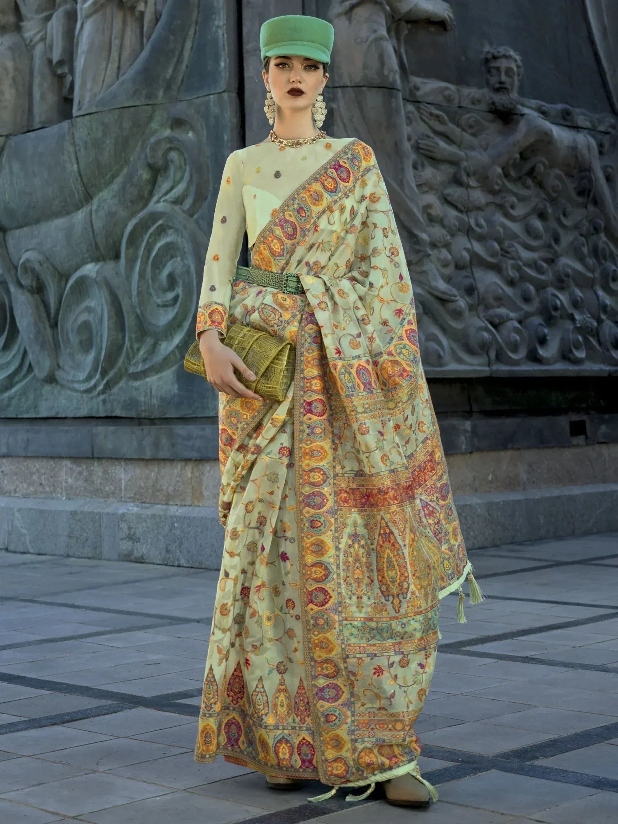 Woman in elegant printed saree with green cap, ethnic jewelry, and clutch, styled for festive occasions.