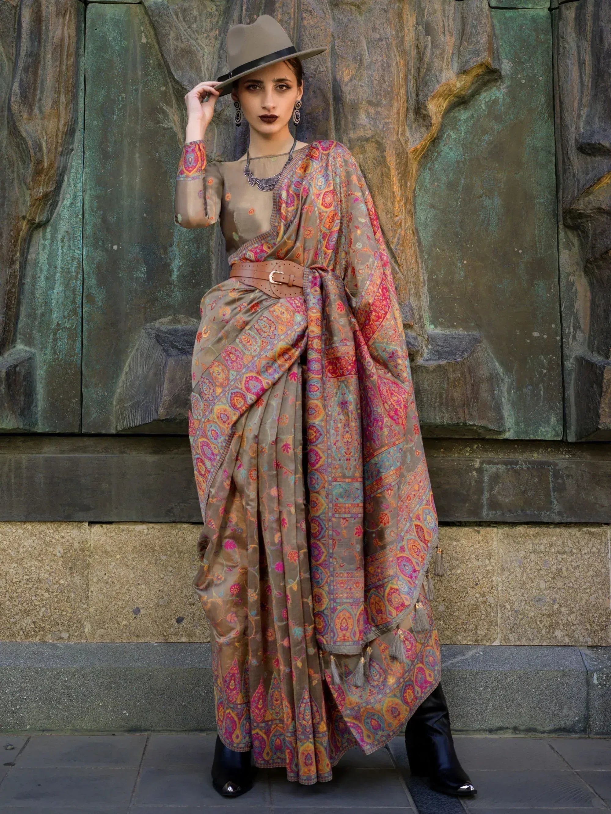Woman in ethnic printed saree with belt, hat, and boots against stone wall