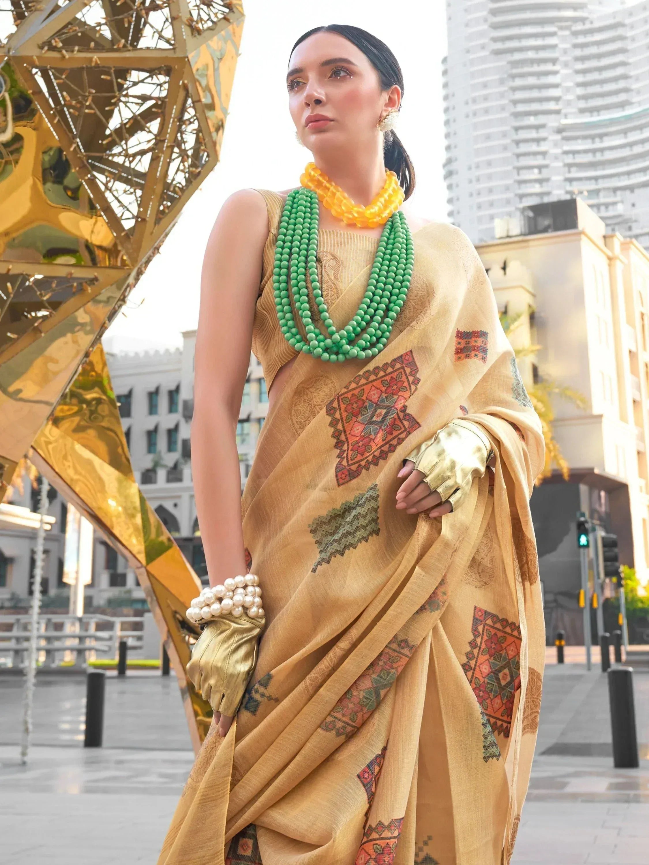 Woman in mustard yellow printed saree with green and yellow beaded necklaces, gold gloves, urban backdrop