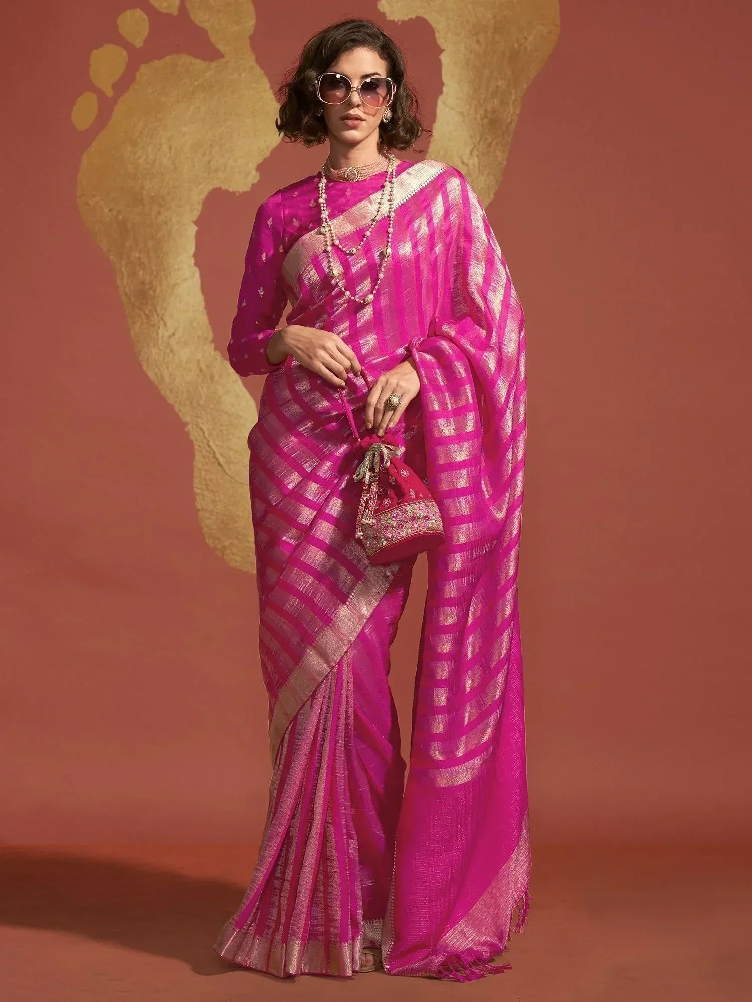 Vibrant pink ethnic saree with gold stripes, paired with matching blouse and potli bag, styled for festive occasions.