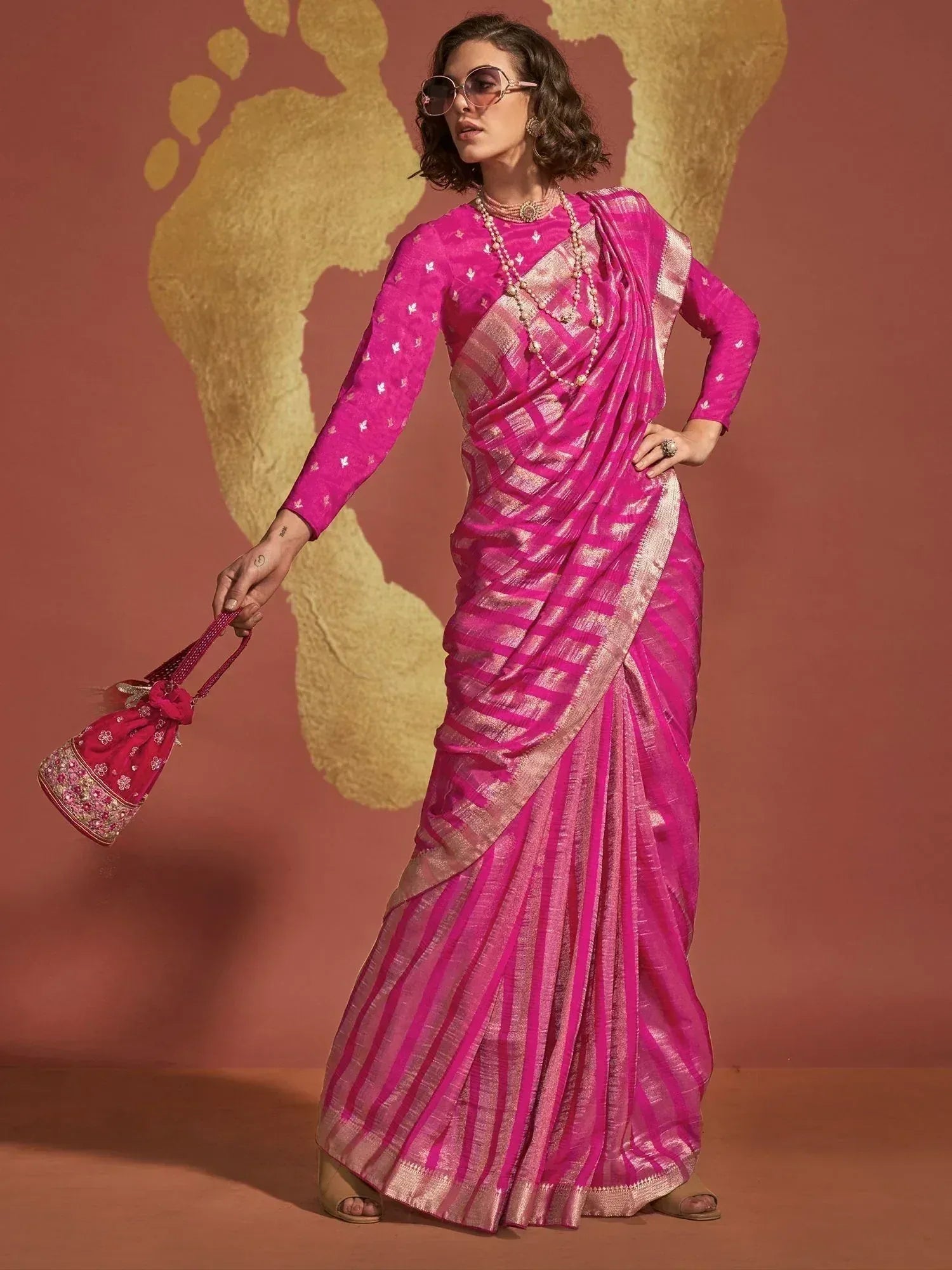 Vibrant pink ethnic saree with gold stripes, paired with matching blouse and potli bag, perfect for Indian occasions.