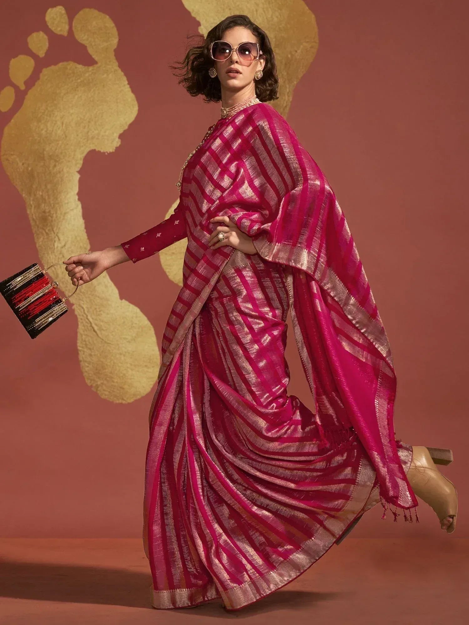 Woman in vibrant pink striped saree with gold border, holding clutch, stylish ethnic wear