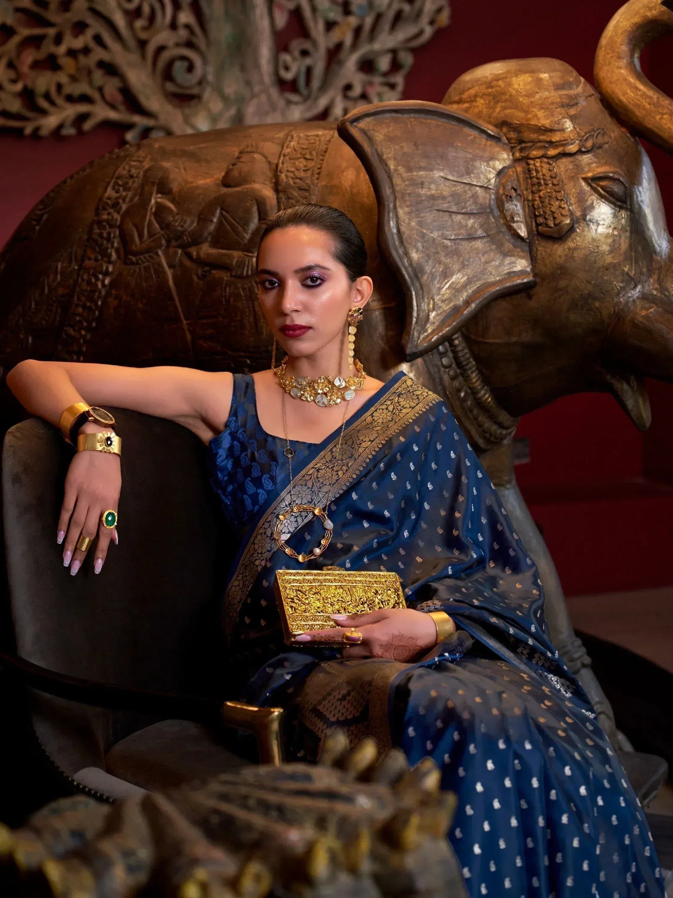 Woman in elegant blue Indian saree with gold jewelry, seated on carved elephant chair, showcasing ethnic fashion