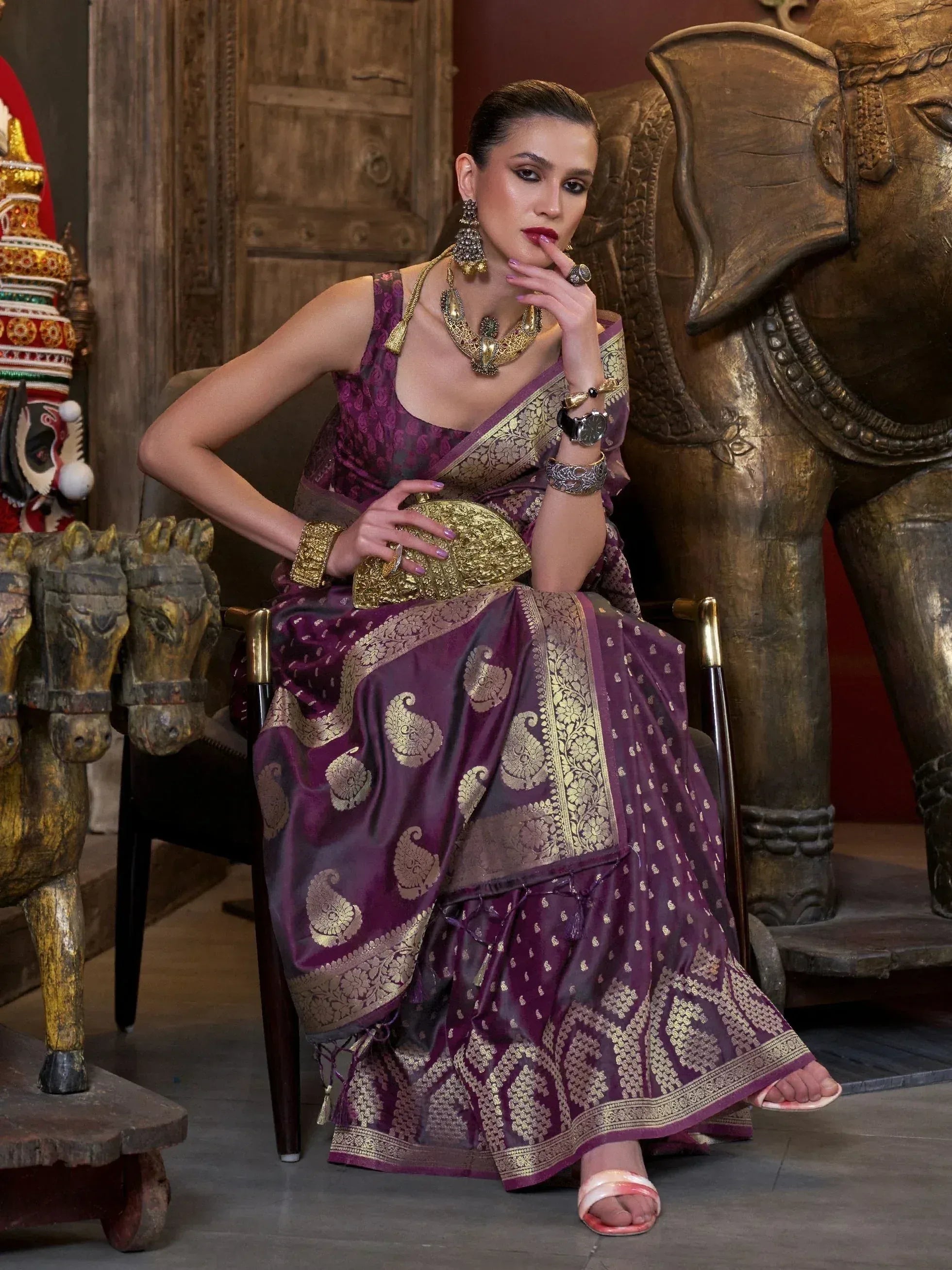 Woman in purple and gold designer saree with intricate paisley patterns, traditional Indian jewelry, and wooden elephant decor.