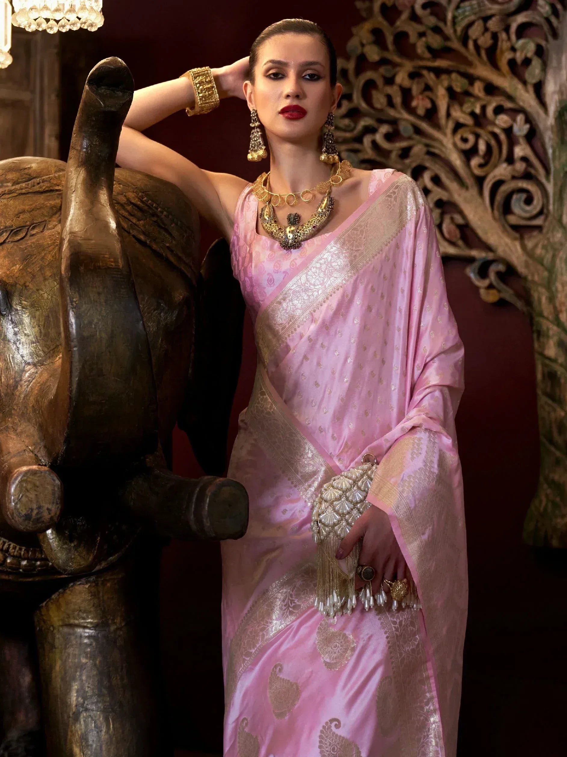 Elegant woman in pink designer saree with gold embroidery, traditional jewelry, and clutch, posing beside carved elephant statue