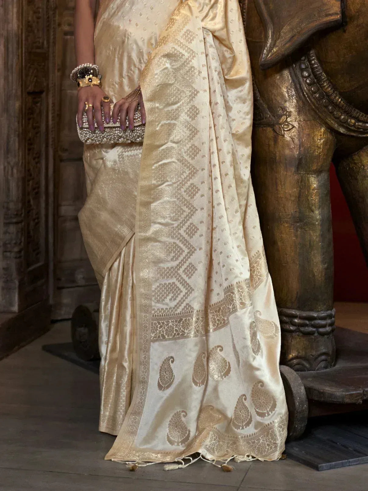 Elegant cream silk saree with gold paisley and geometric motifs, paired with pearl clutch and bangles.