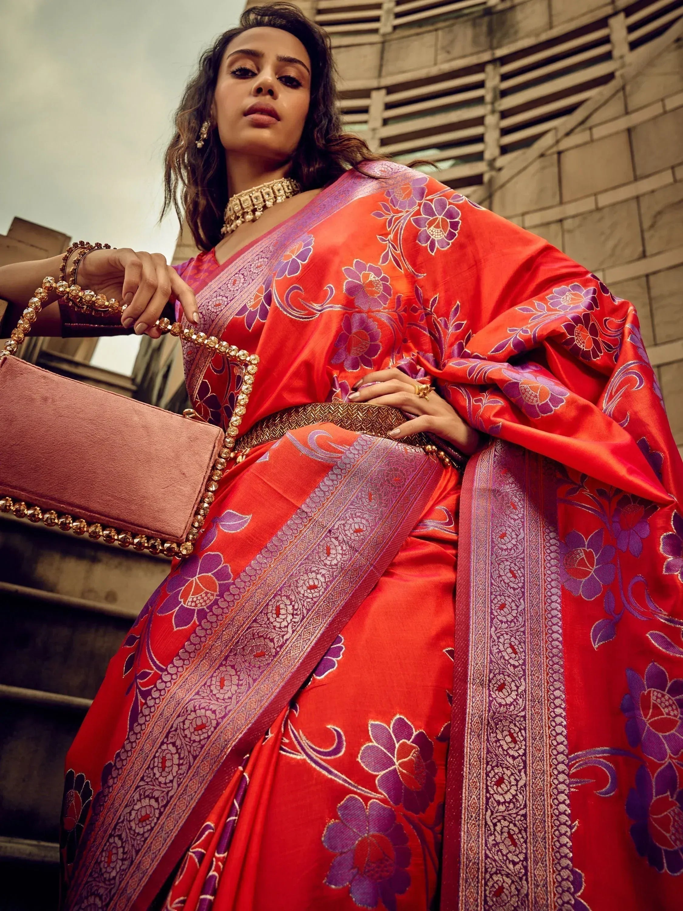 Vibrant red saree with purple floral print and gold border, paired with statement jewelry and pink clutch.
