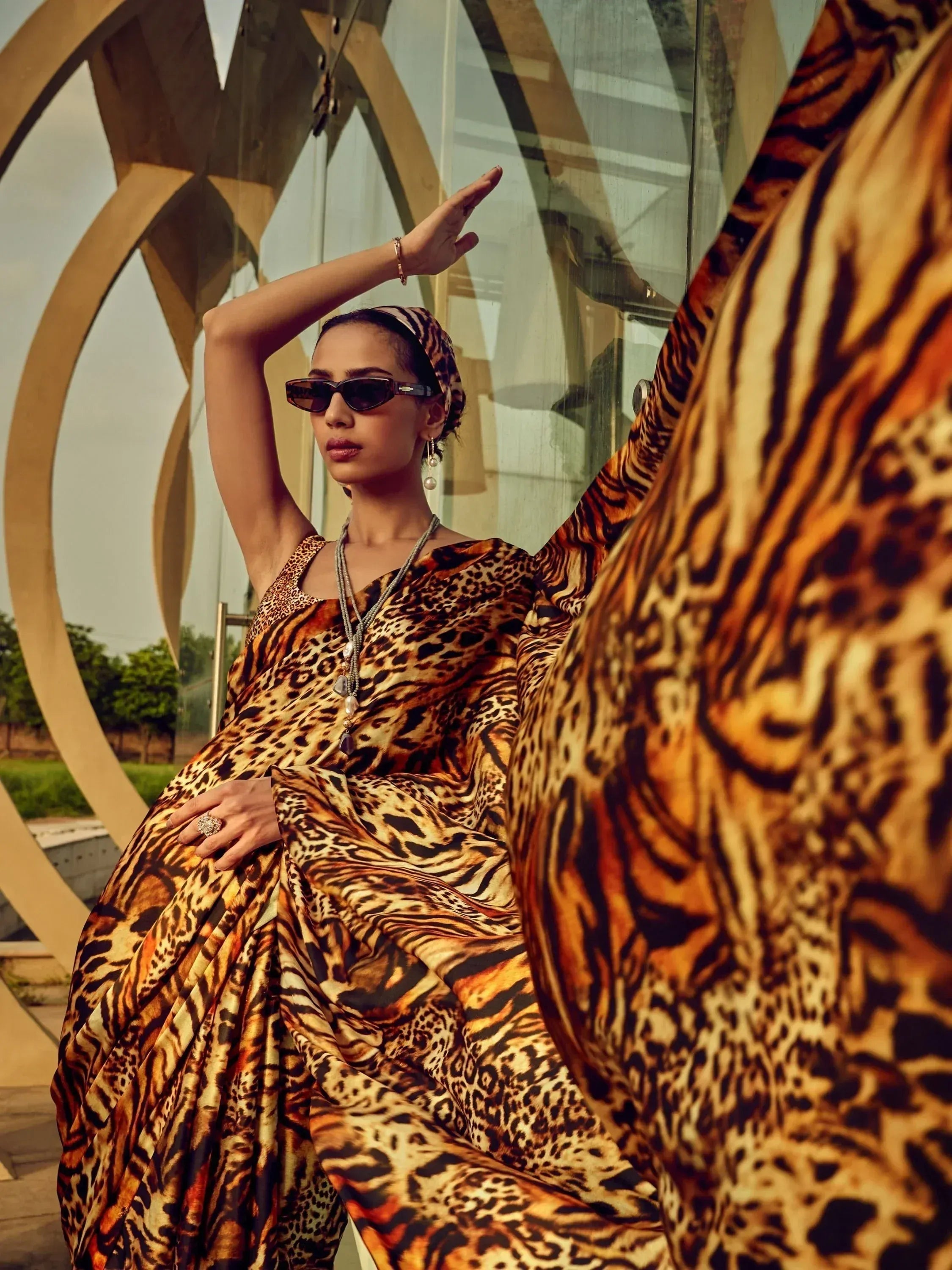 Woman in bold leopard print ethnic gown with matching headscarf, stylish sunglasses, and jewelry, posing outdoors.