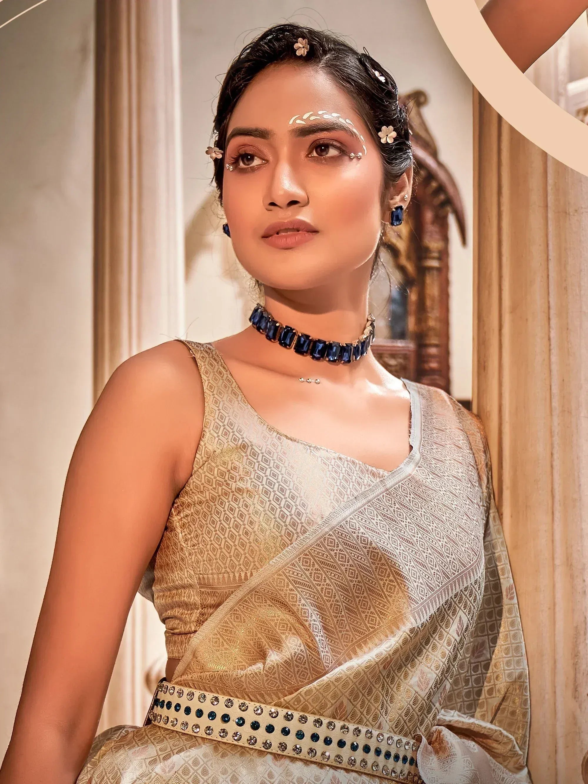 Elegant woman in gold and white patterned saree with blue gem choker, floral hair accessories, and festive makeup.