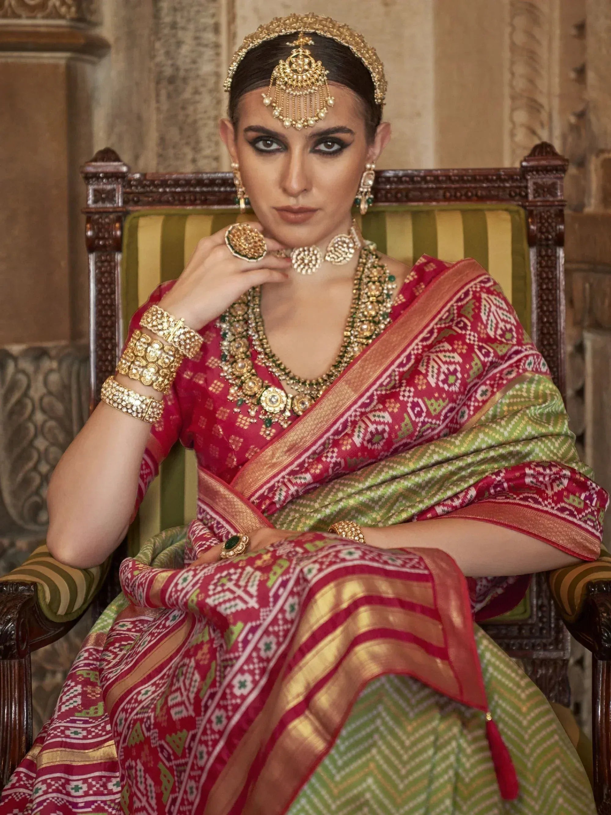 Woman in red and green patterned saree with gold jewelry, seated on ornate chair