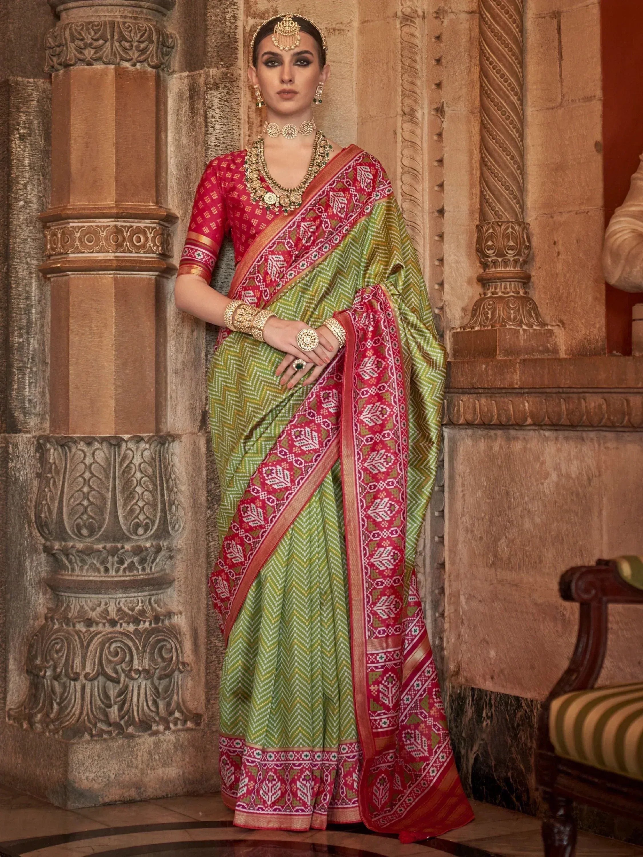 Woman in green and red printed saree with gold jewelry, standing near carved stone pillars