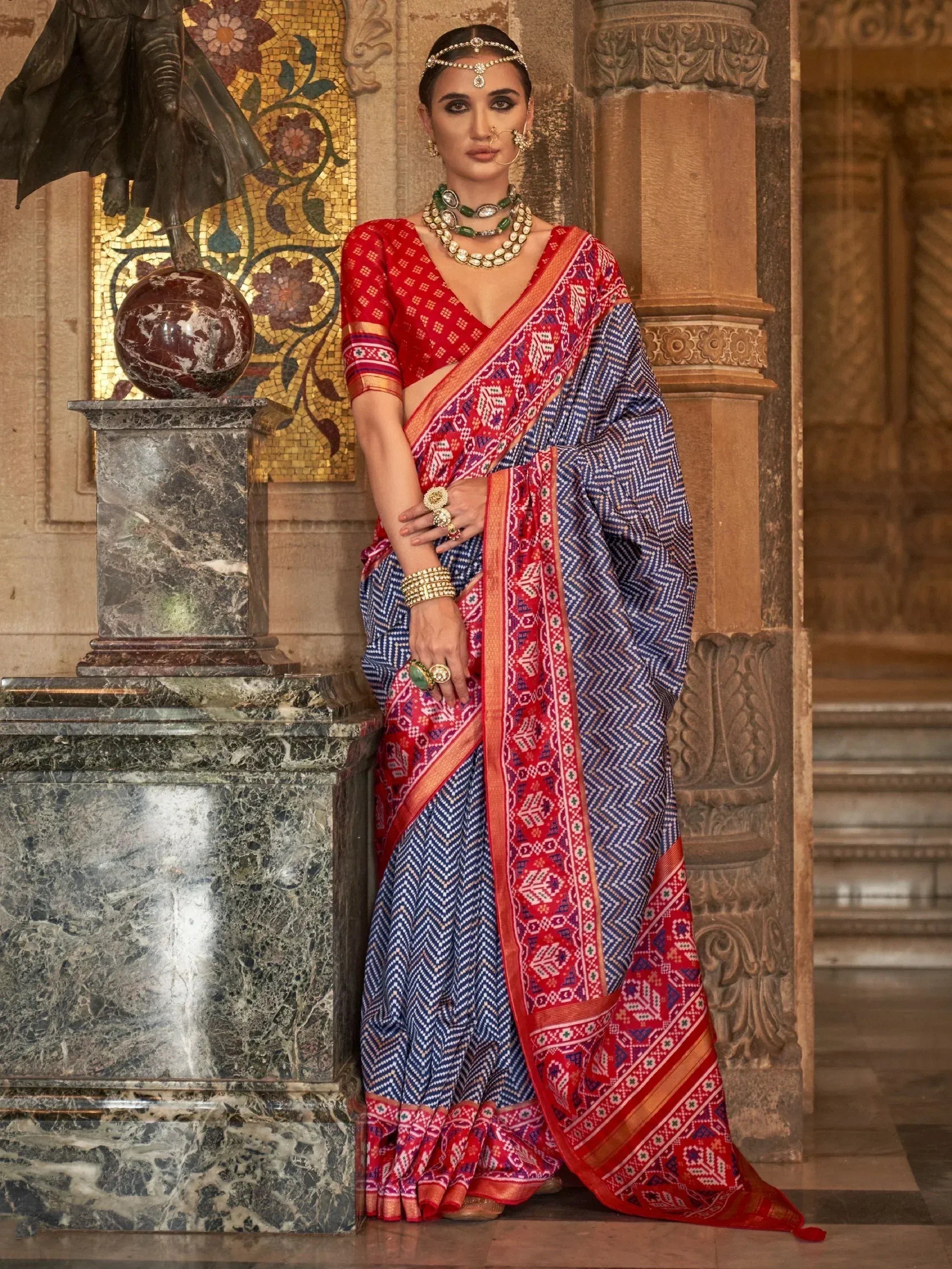 Woman in red and blue ethnic saree with intricate patterns, traditional jewelry, and temple backdrop
