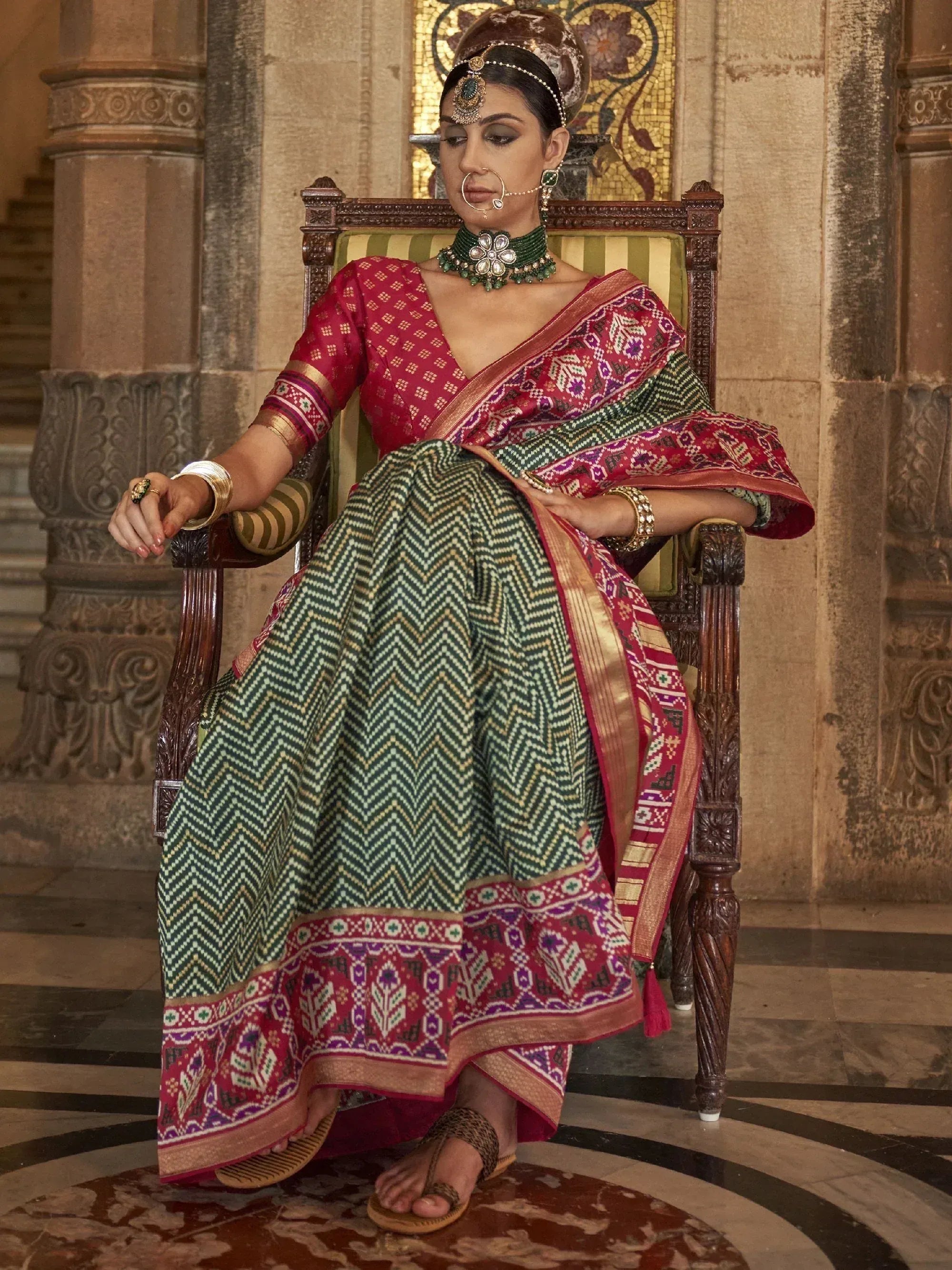 Woman in traditional Indian ethnic wear, red and green patterned lehenga choli with intricate jewelry, seated on ornate chair