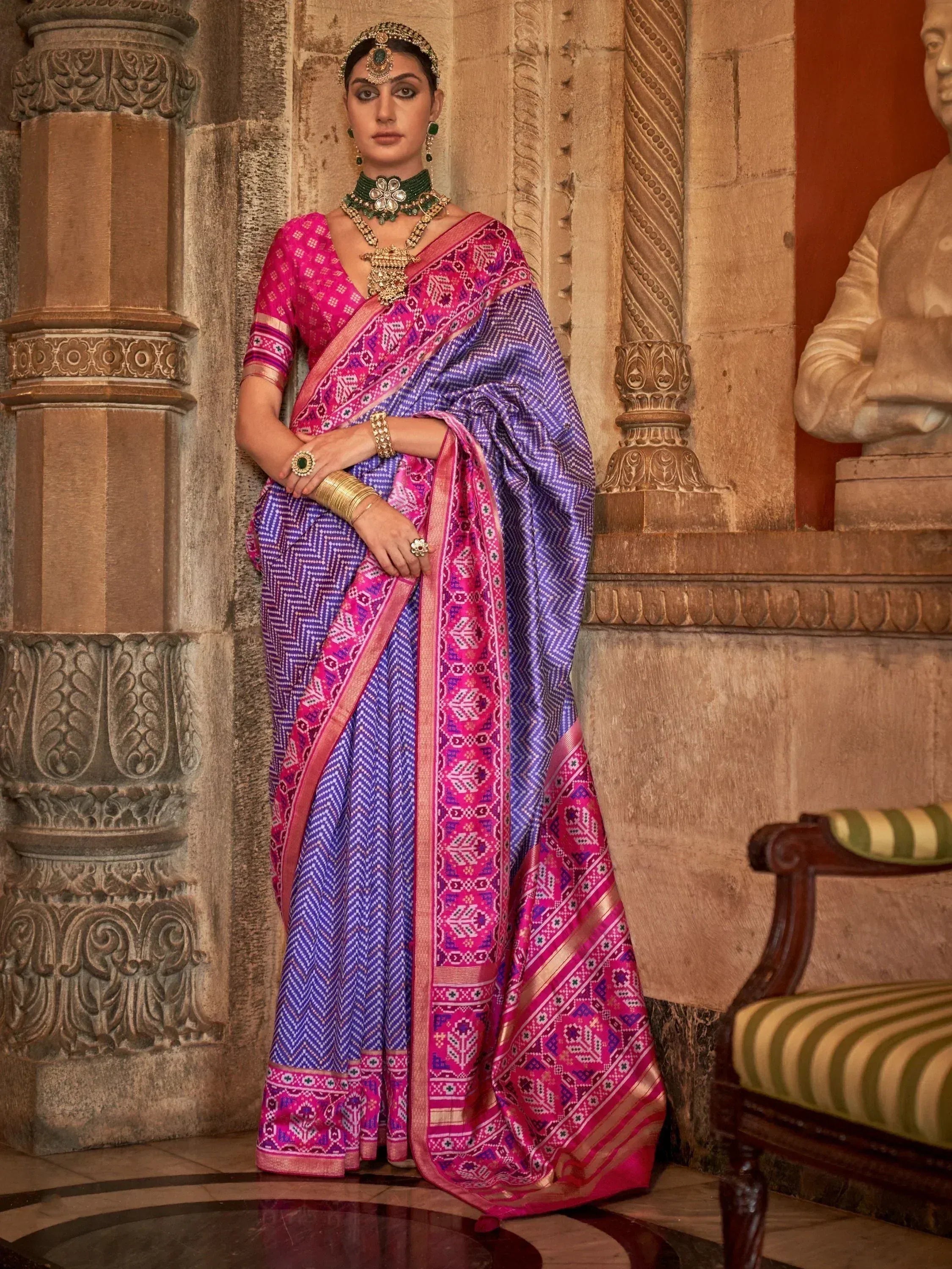Woman in vibrant purple and pink ethnic saree with intricate patterns, traditional jewelry, and ornate backdrop