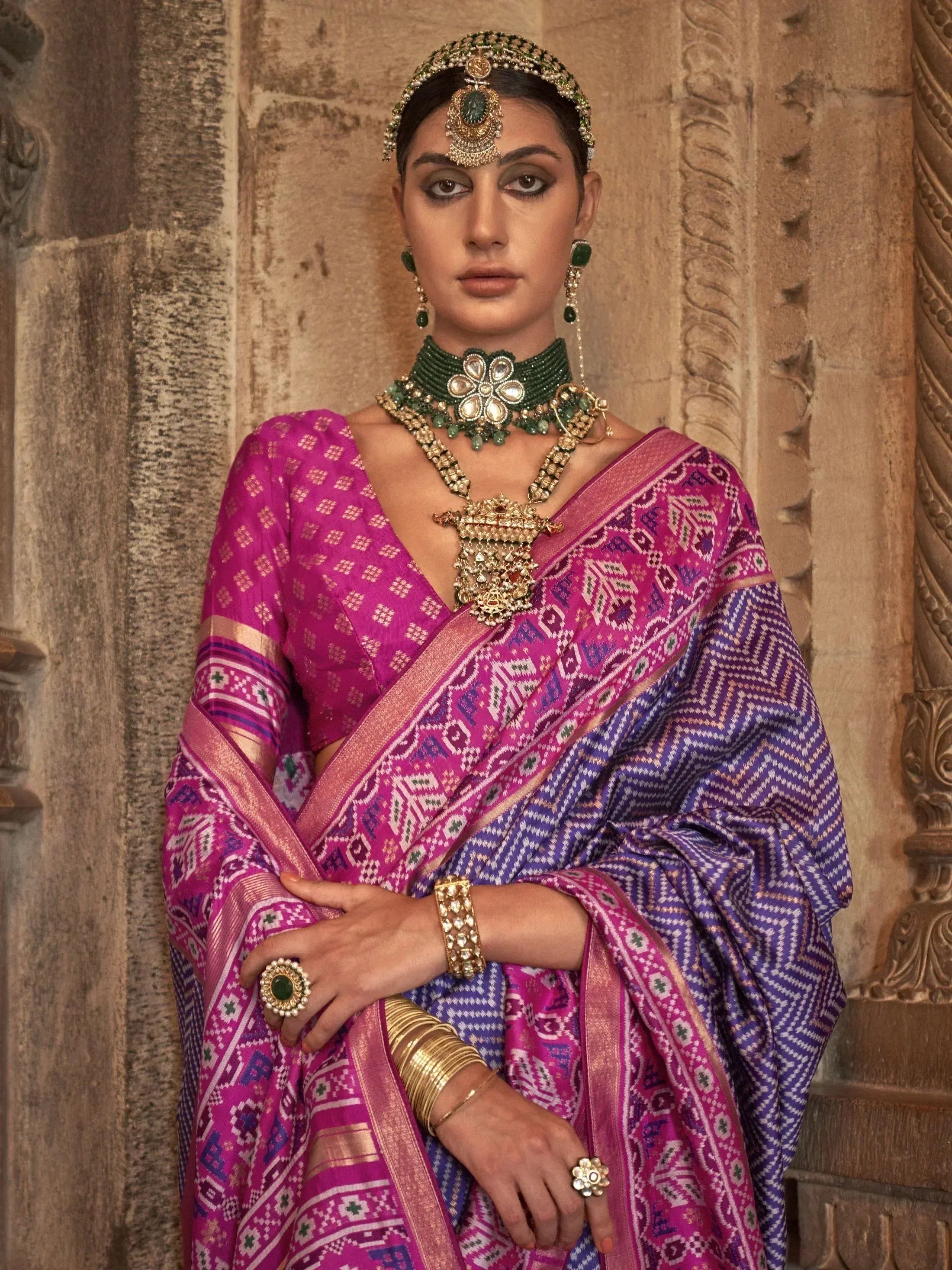 Woman in vibrant pink and purple ethnic saree with intricate patterns, traditional jewelry, and temple backdrop