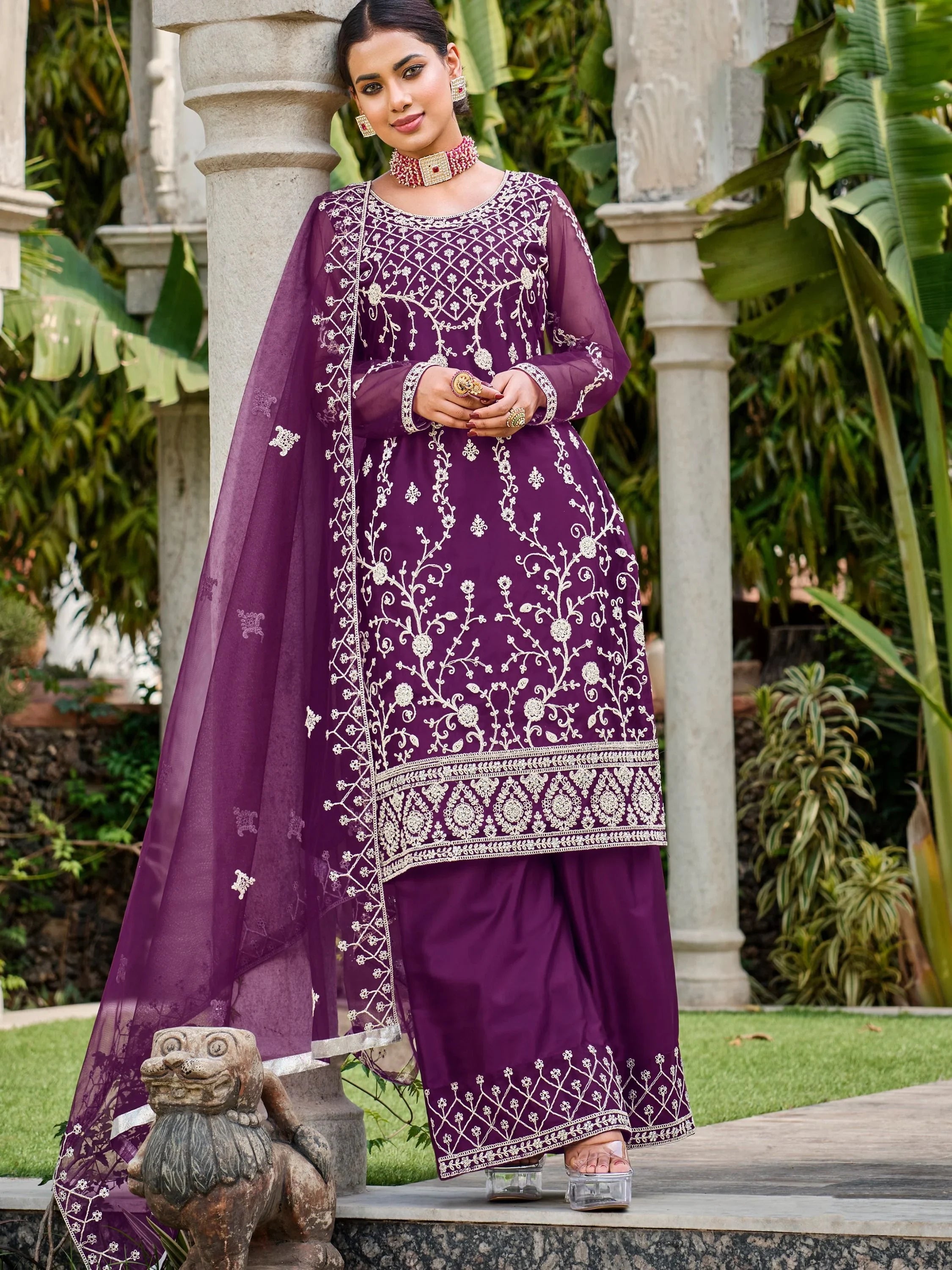 Woman in a purple embroidered salwar kameez and dupatta, posing outdoors in a garden setting