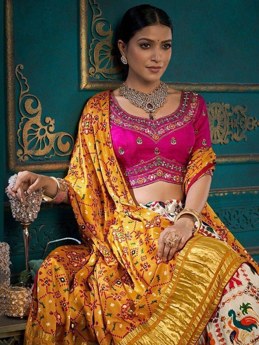 Woman in vibrant pink and gold embroidered lehenga choli with traditional jewelry, seated against ornate teal backdrop