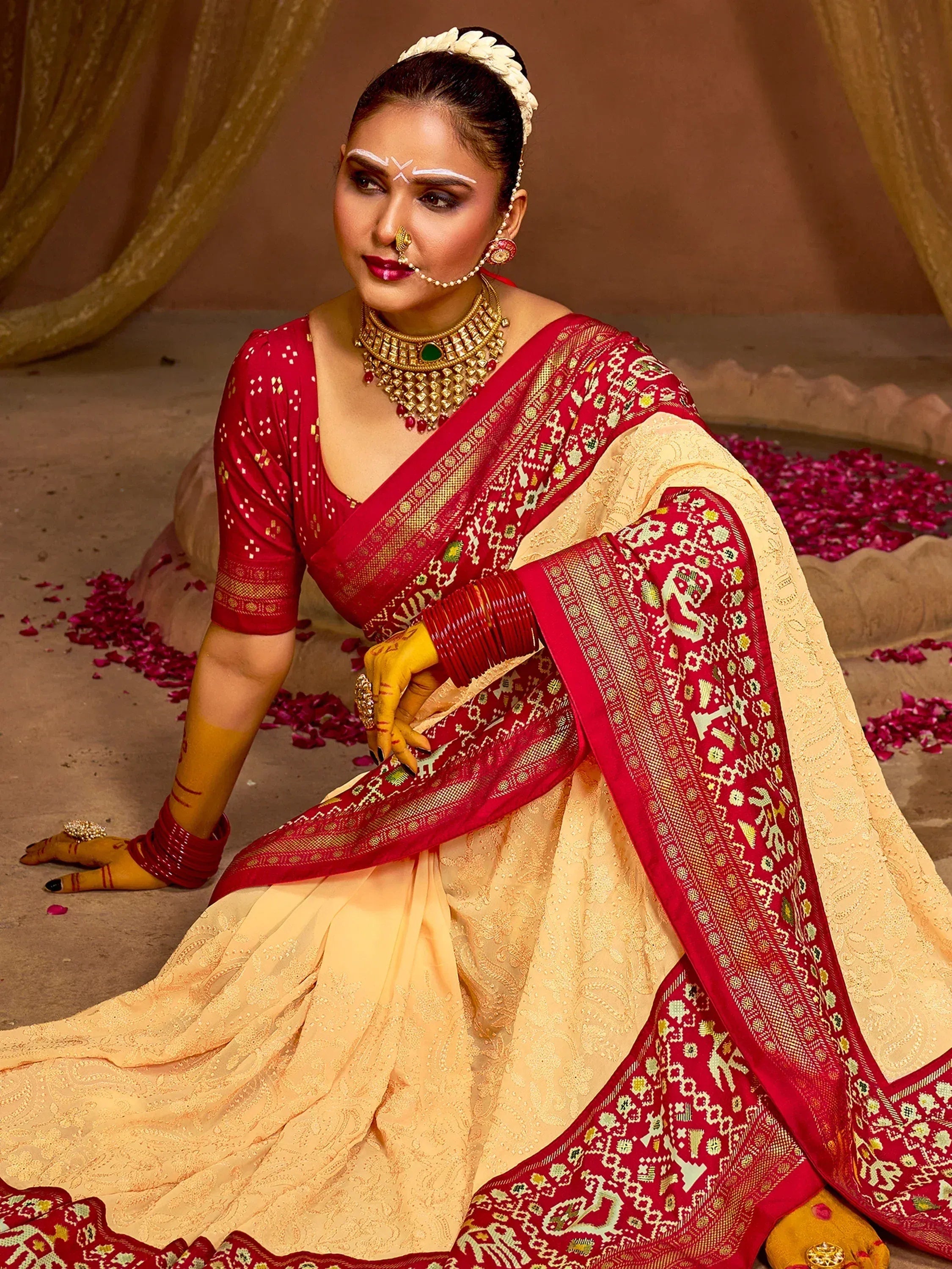 Woman in red and cream ethnic saree with gold jewelry, henna hands, and floral backdrop