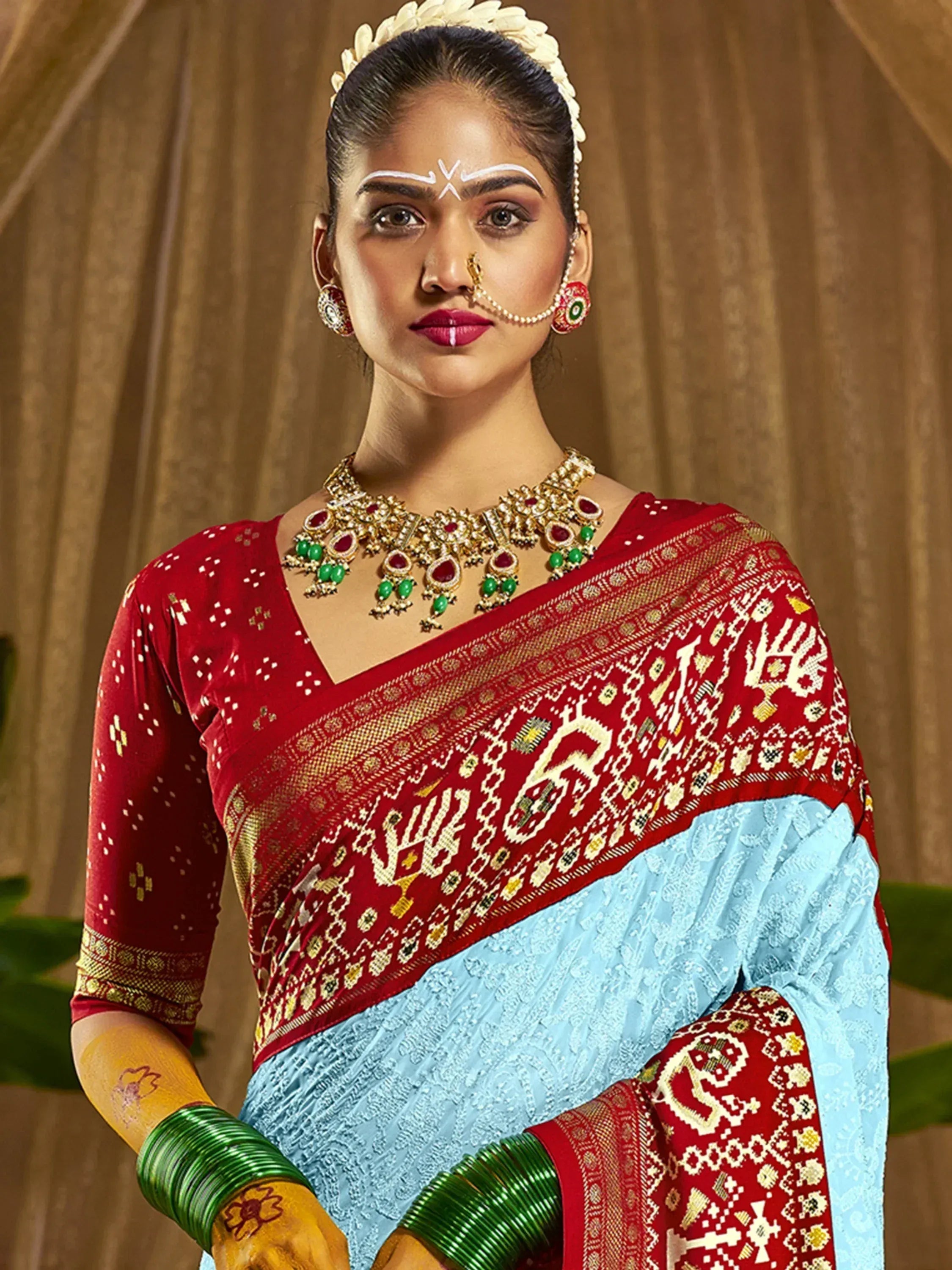 Red and blue ethnic saree with gold embroidery, traditional jewelry, and floral headpiece for Indian occasions