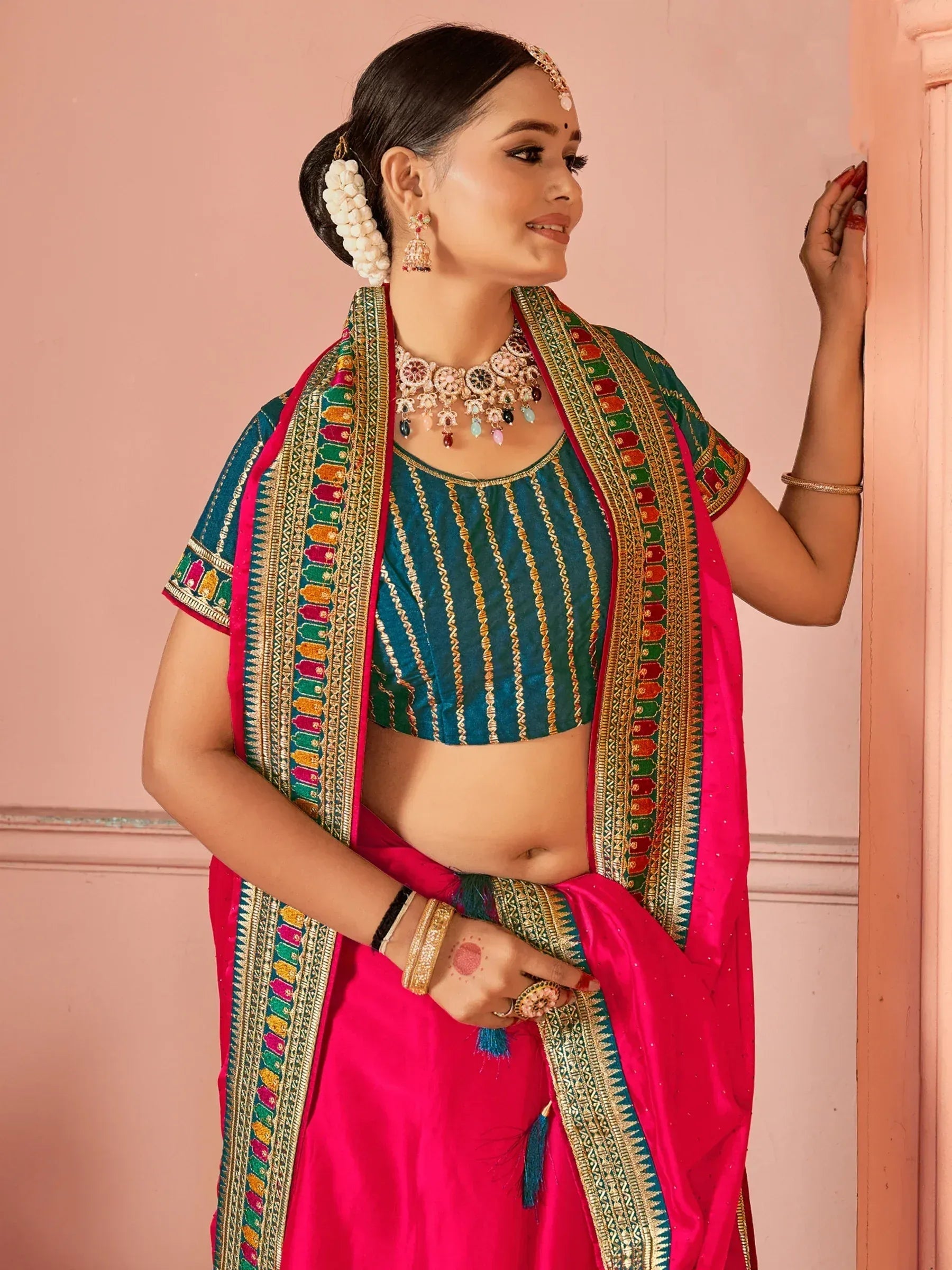 Woman in vibrant pink and teal Indian ethnic saree with gold embroidery, traditional jewelry, and floral hair accessory