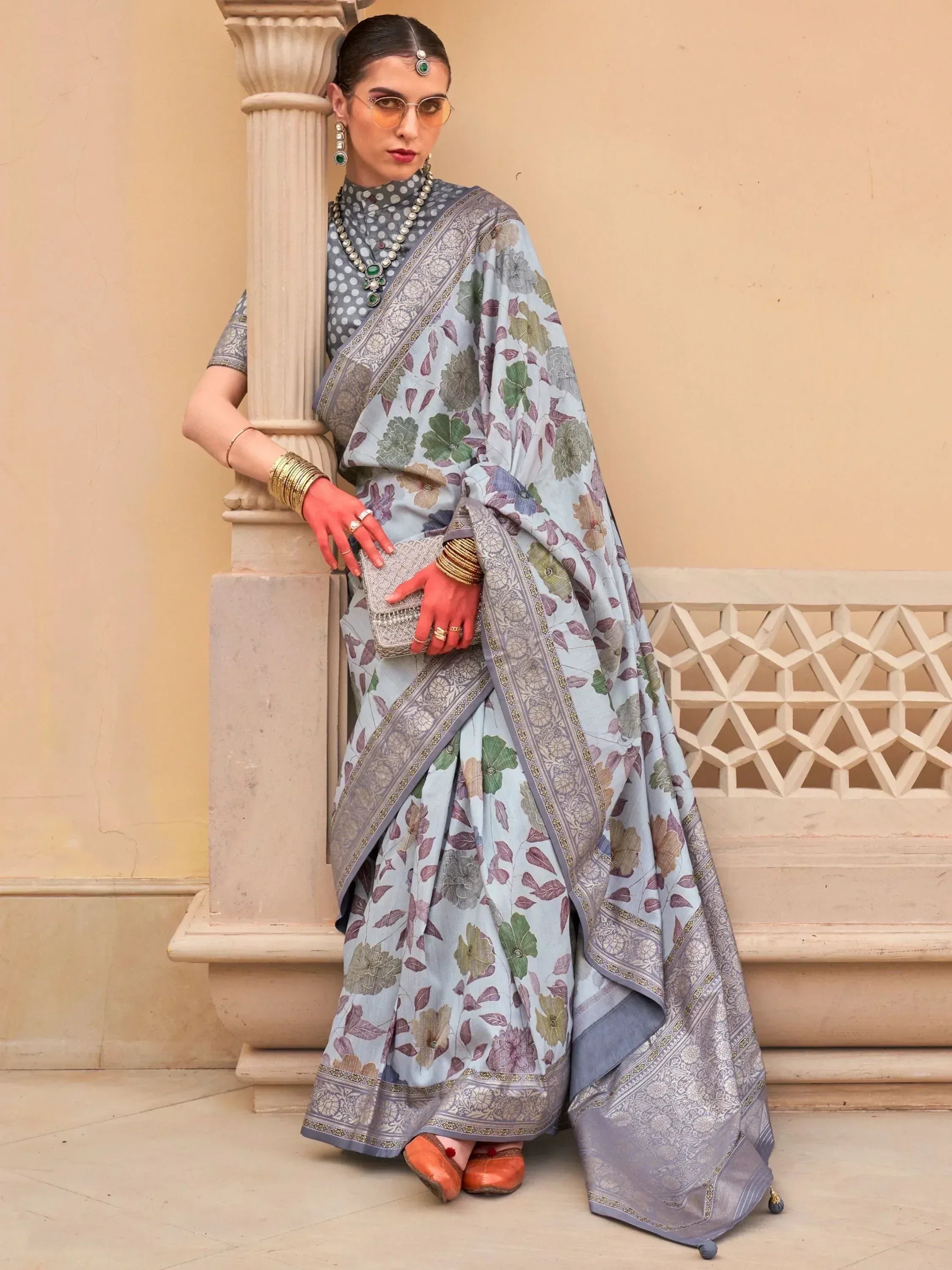 Woman in floral print saree with silver border, ethnic jewelry, and orange flats, leaning on pillar