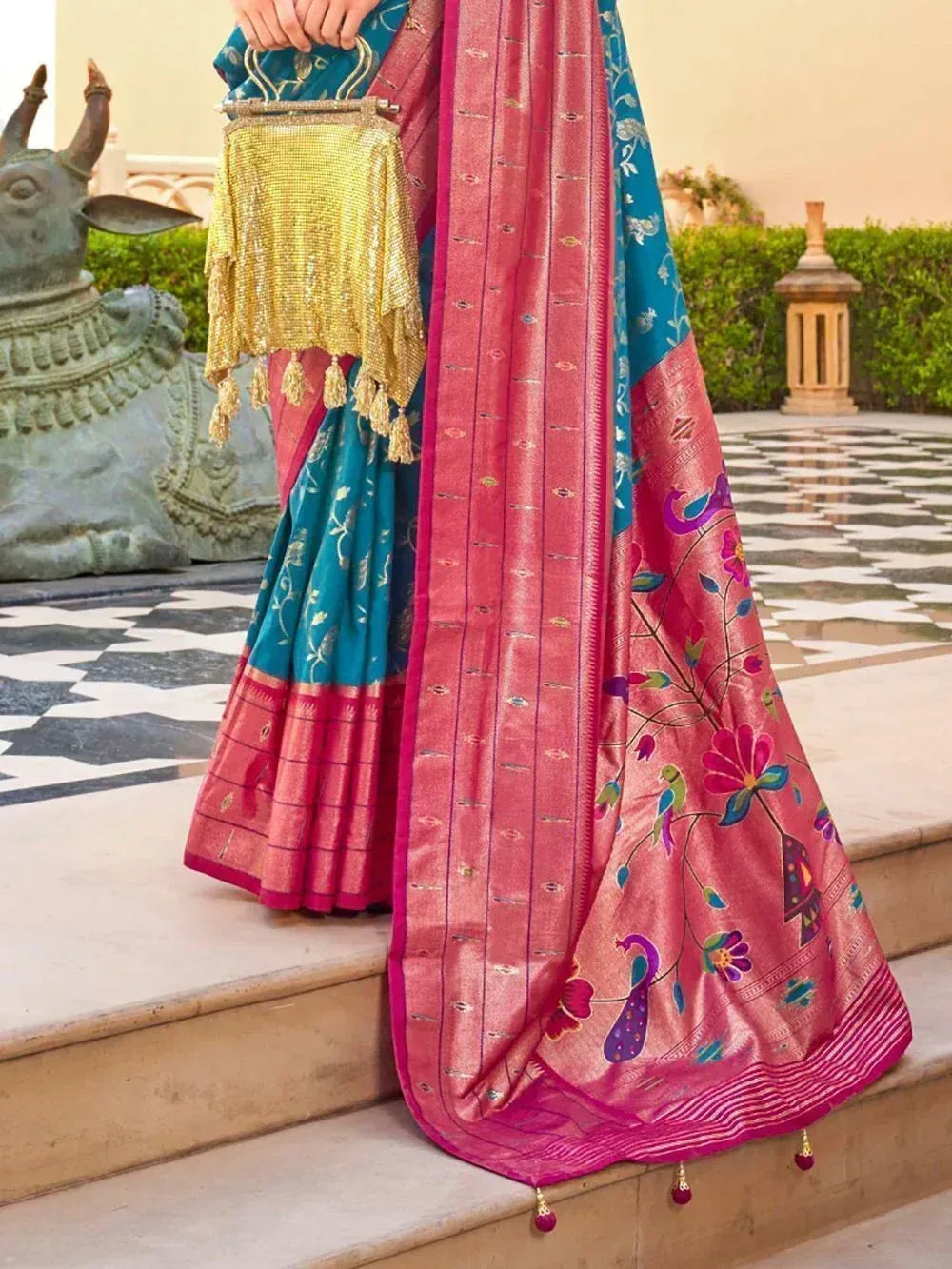 Pink and teal designer saree with peacock floral embroidery, paired with gold embellished handbag, perfect for Indian ethnic occasions.
