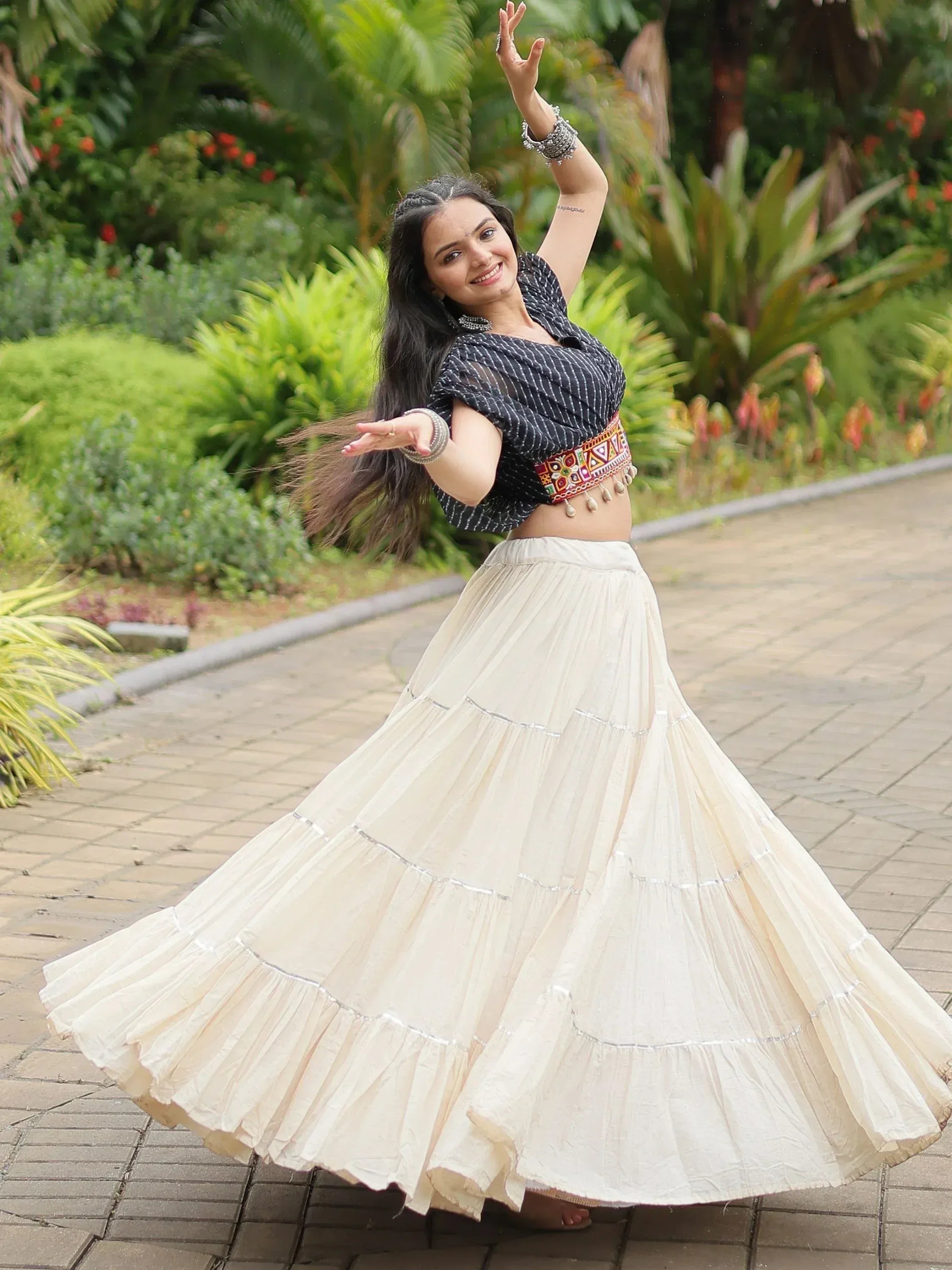 Woman dancing in black printed crop top and cream tiered ethnic skirt with embroidered belt, garden setting