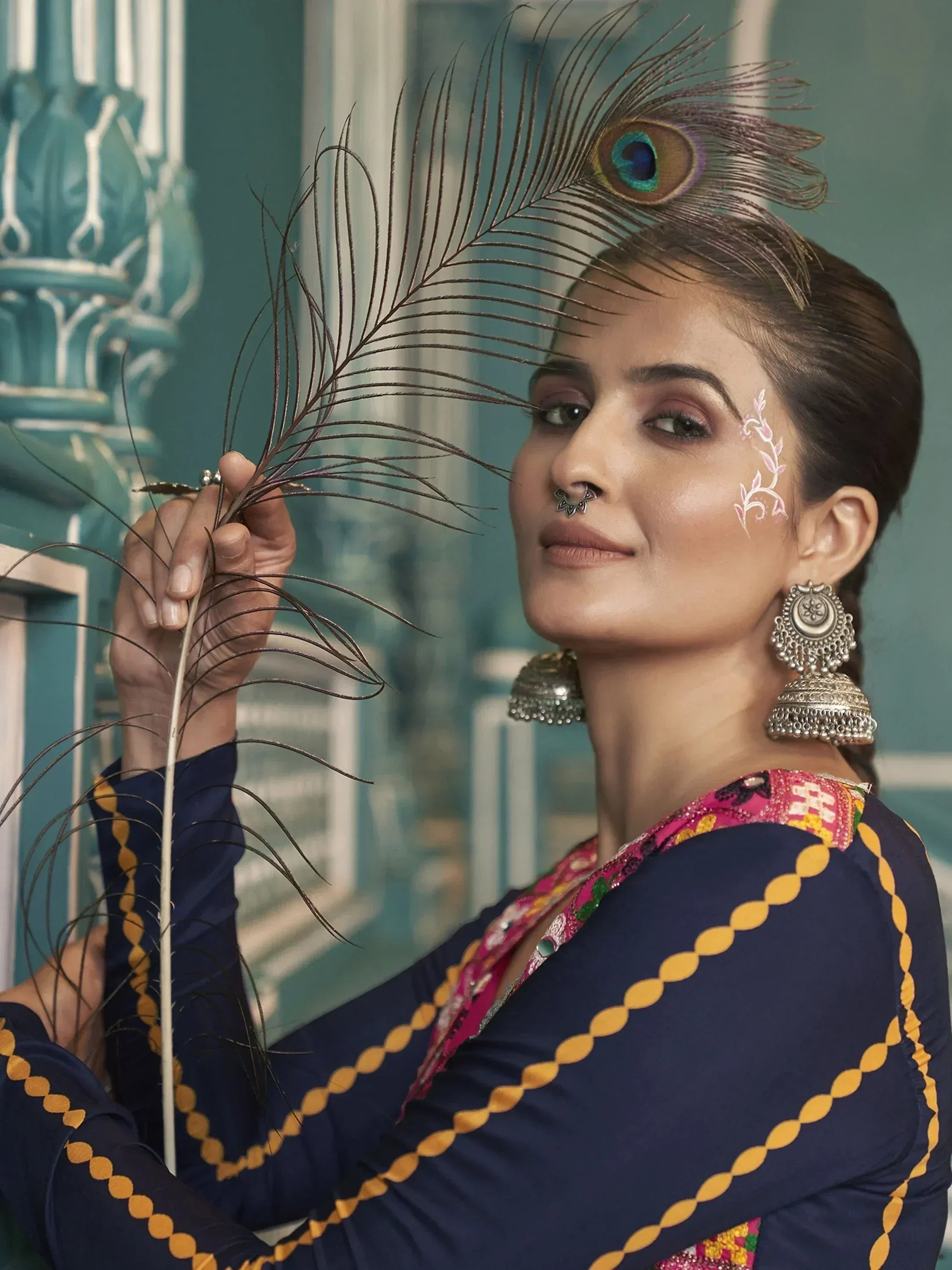 Woman in ethnic navy blue outfit with peacock feather, traditional jewelry, and floral face art