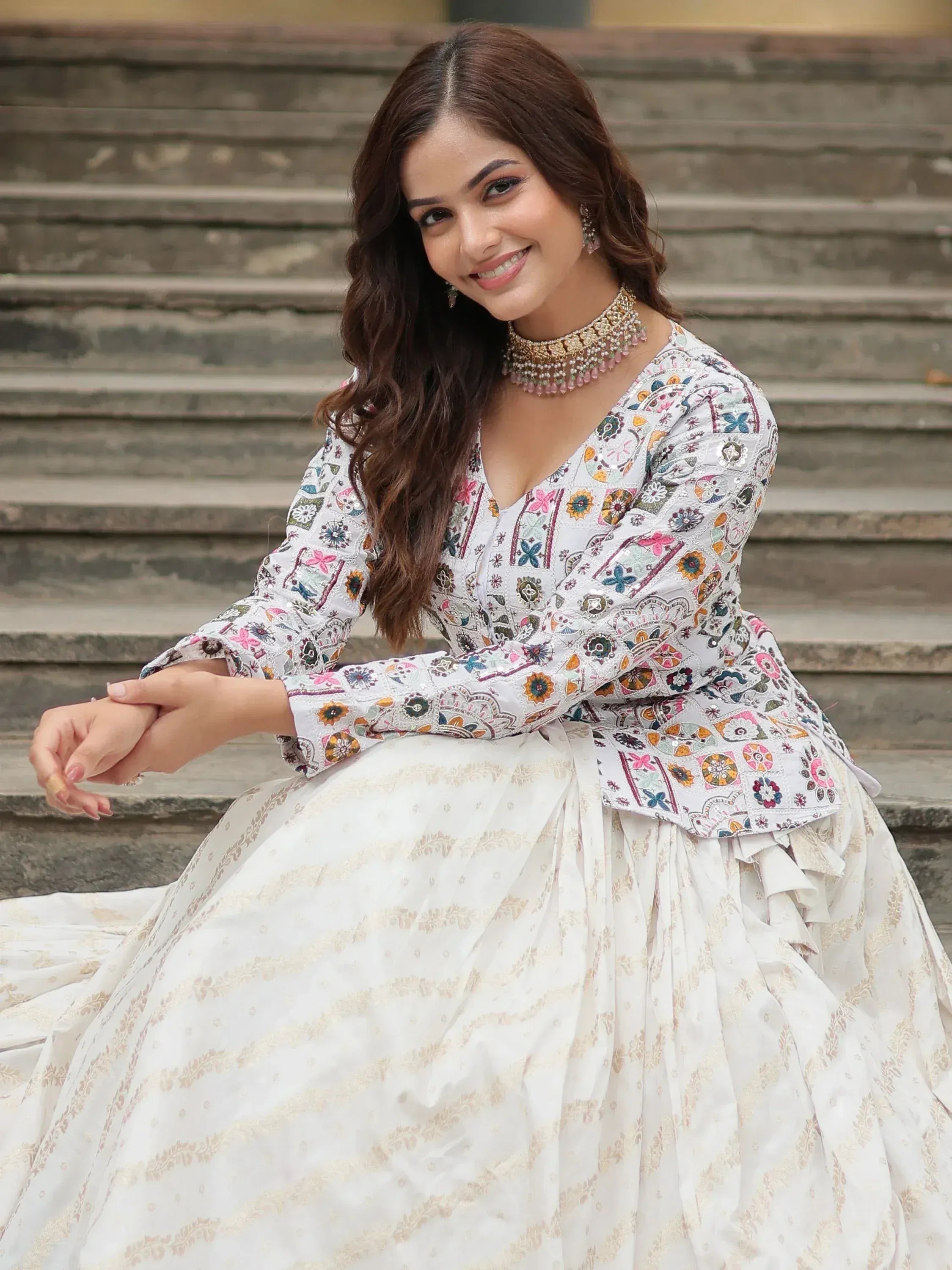 Woman in white floral embroidered lehenga choli with gold detailing, sitting on stone steps