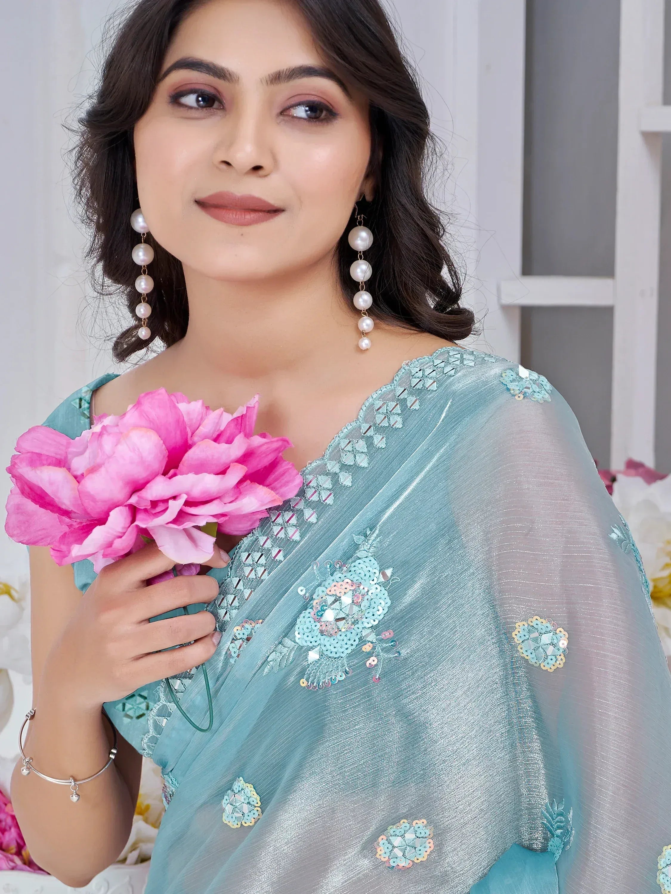 Woman in light blue embroidered saree with pearl earrings, holding pink flower, Indian ethnic wear
