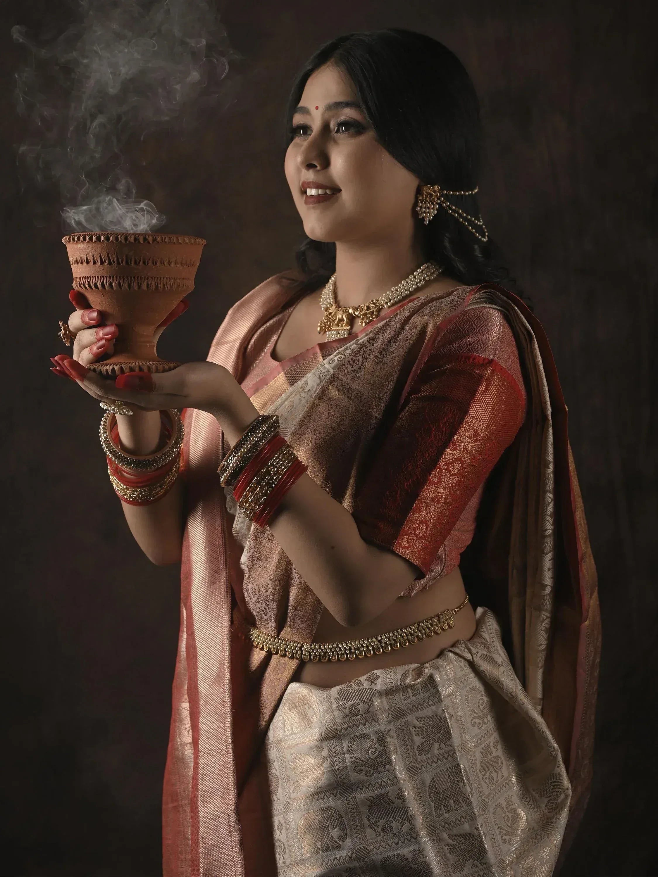 Woman in traditional Indian saree holding smoking clay pot, adorned with gold jewelry and bangles