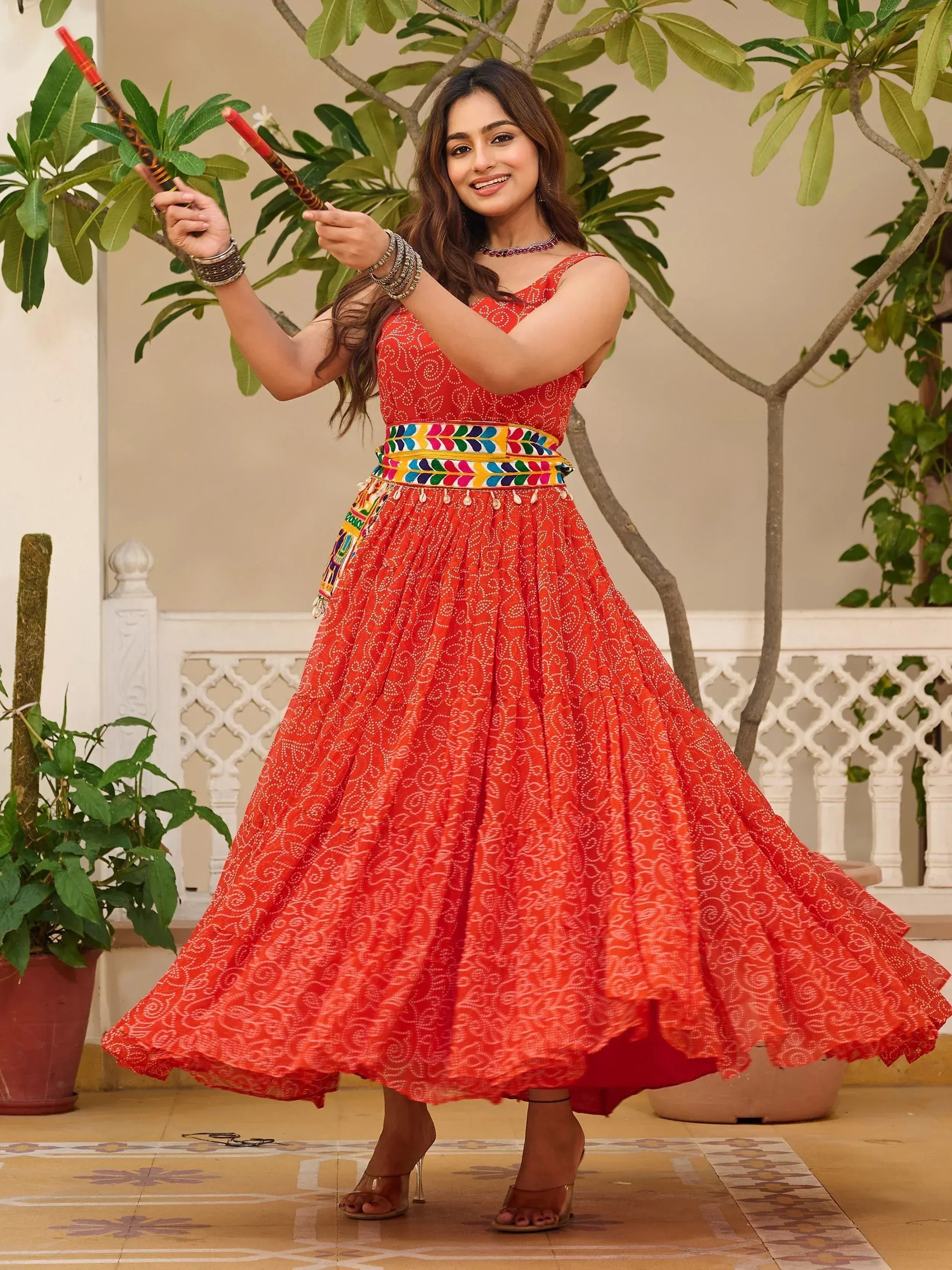 Woman in vibrant red ethnic gown with printed patterns, colorful belt, and dandiya sticks, perfect for festive occasions.