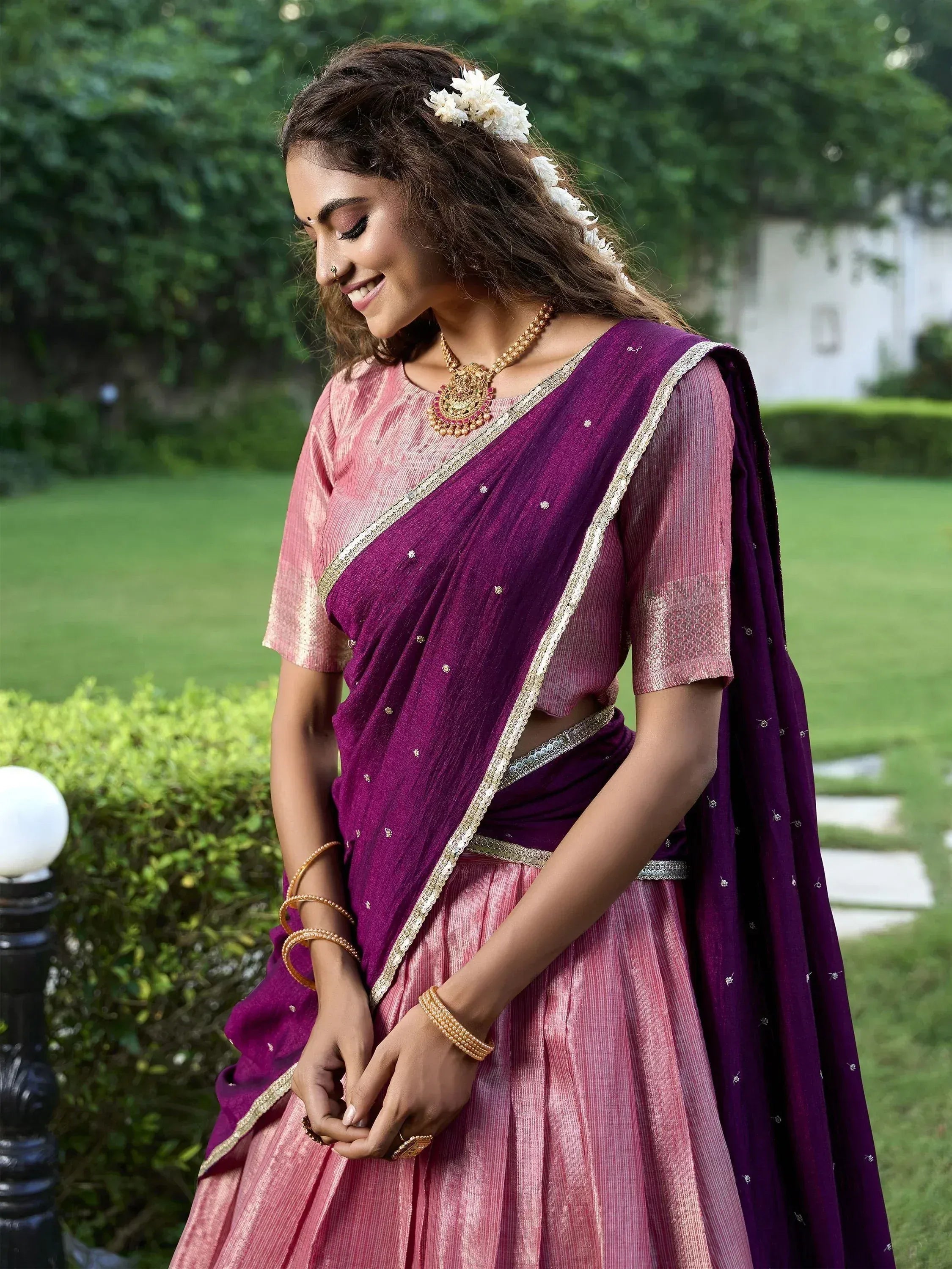 Woman in purple and pink designer lehenga choli with gold embroidery, floral hair accessory, and traditional jewelry