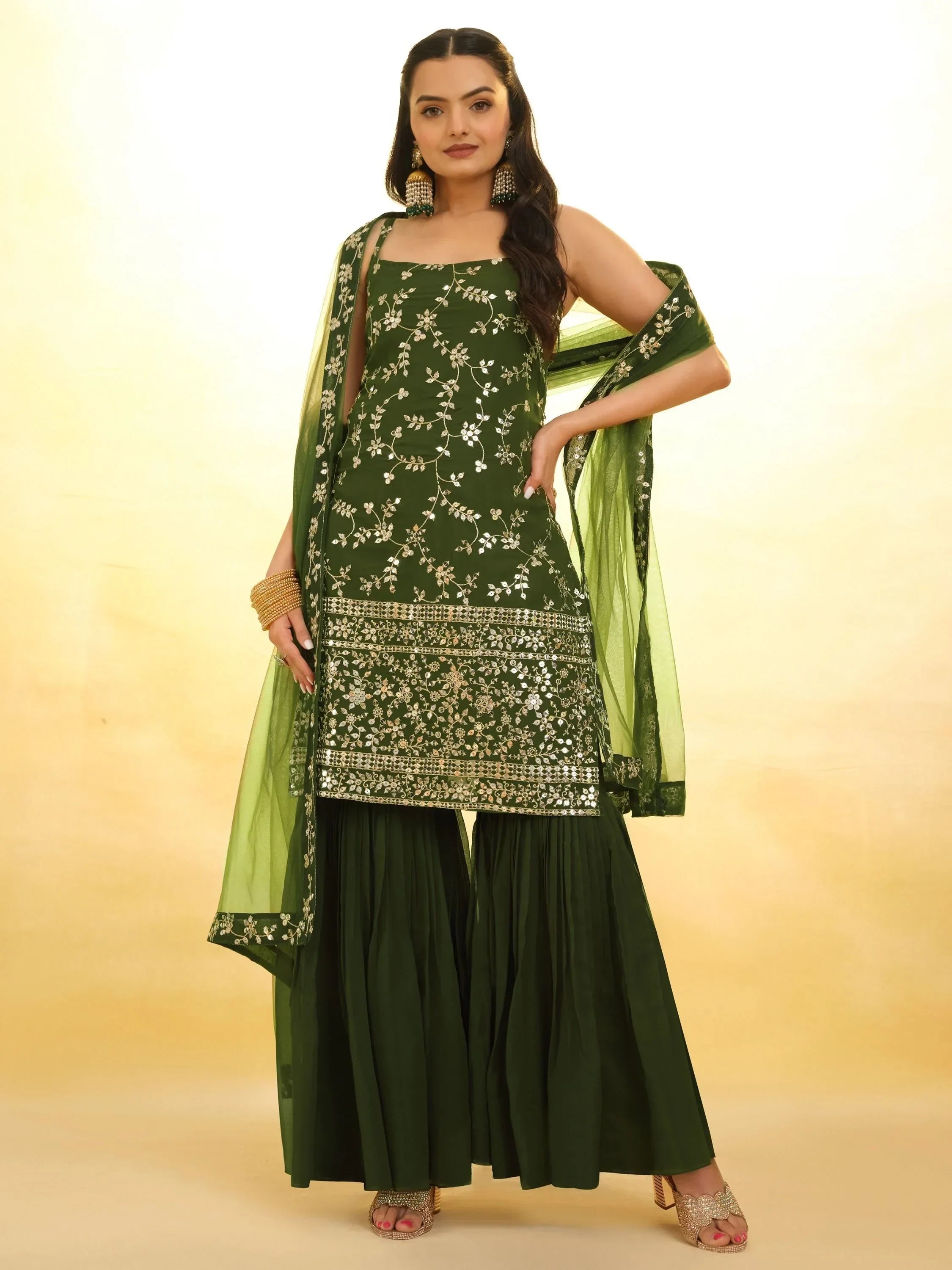 Woman wearing a dark green embroidered ethnic sharara suit with dupatta on light background