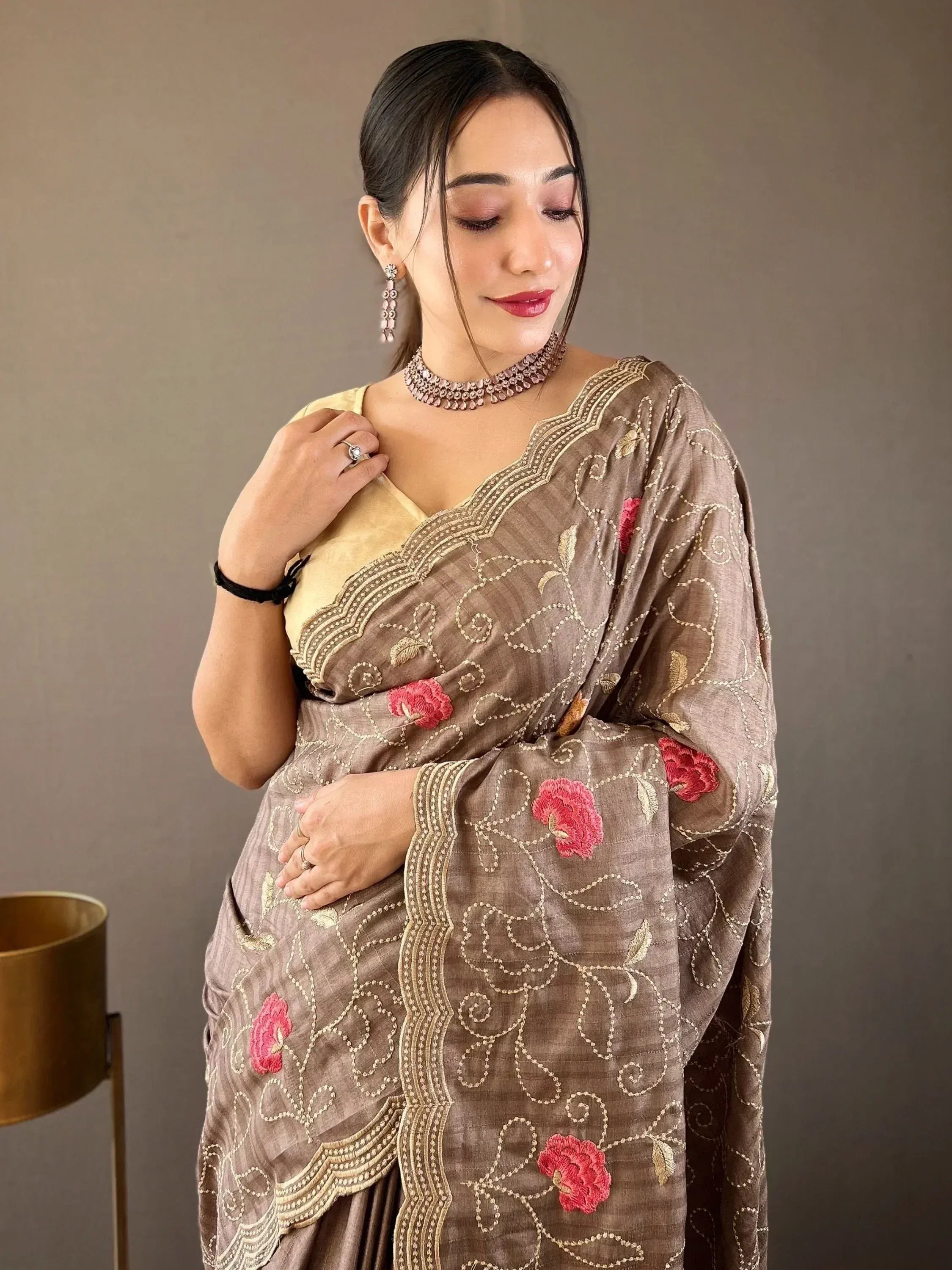Woman in elegant brown saree with pink floral embroidery and gold detailing, paired with traditional jewelry